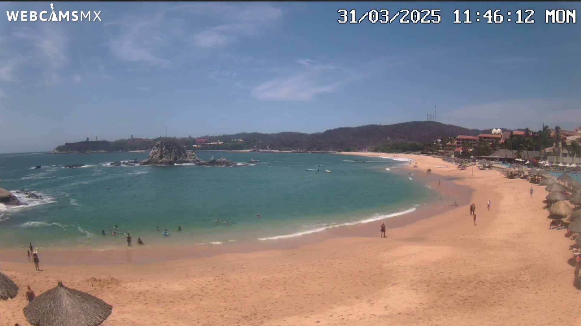 A sandy beach with calm, clear turquoise water curves around a rocky outcrop under a mostly sunny sky.