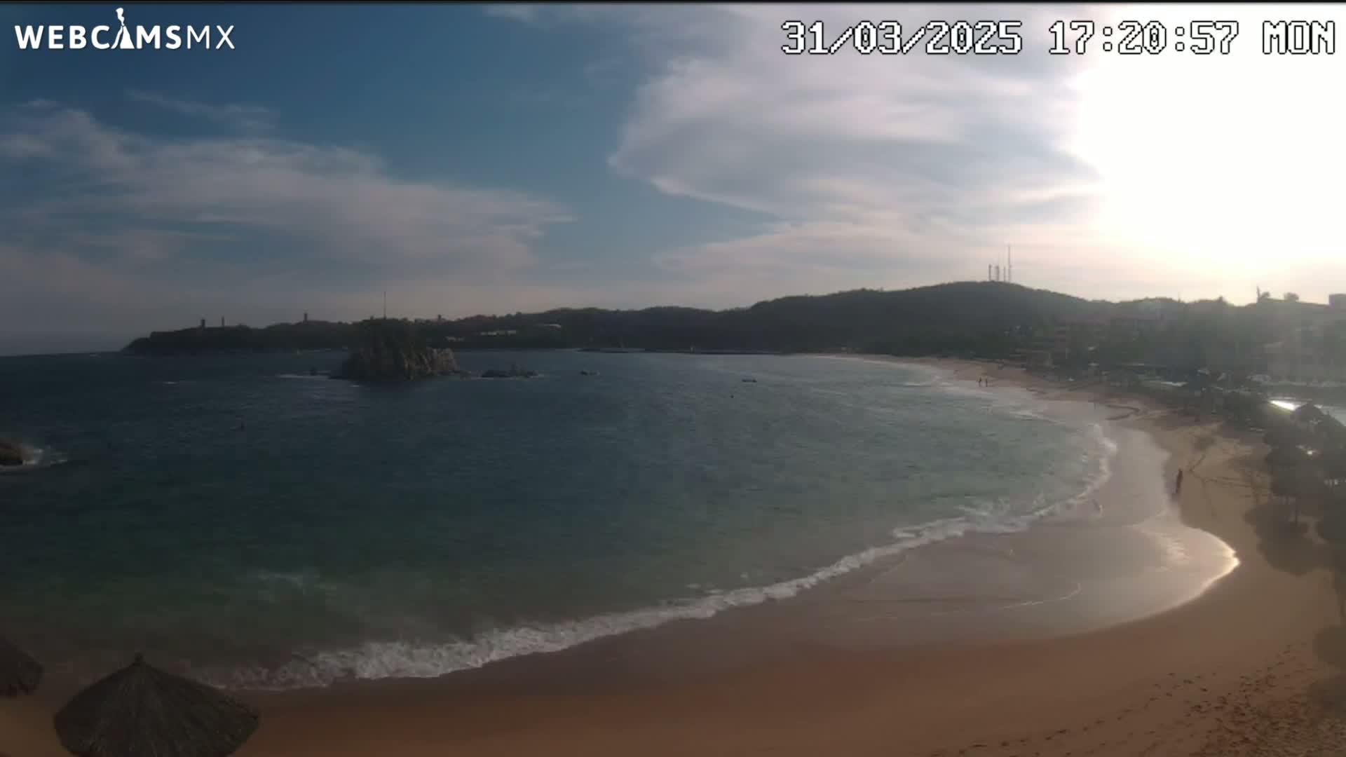 A partly cloudy day shows a sandy beach with gentle waves lapping the shore, a calm bay, and a rocky island offshore.