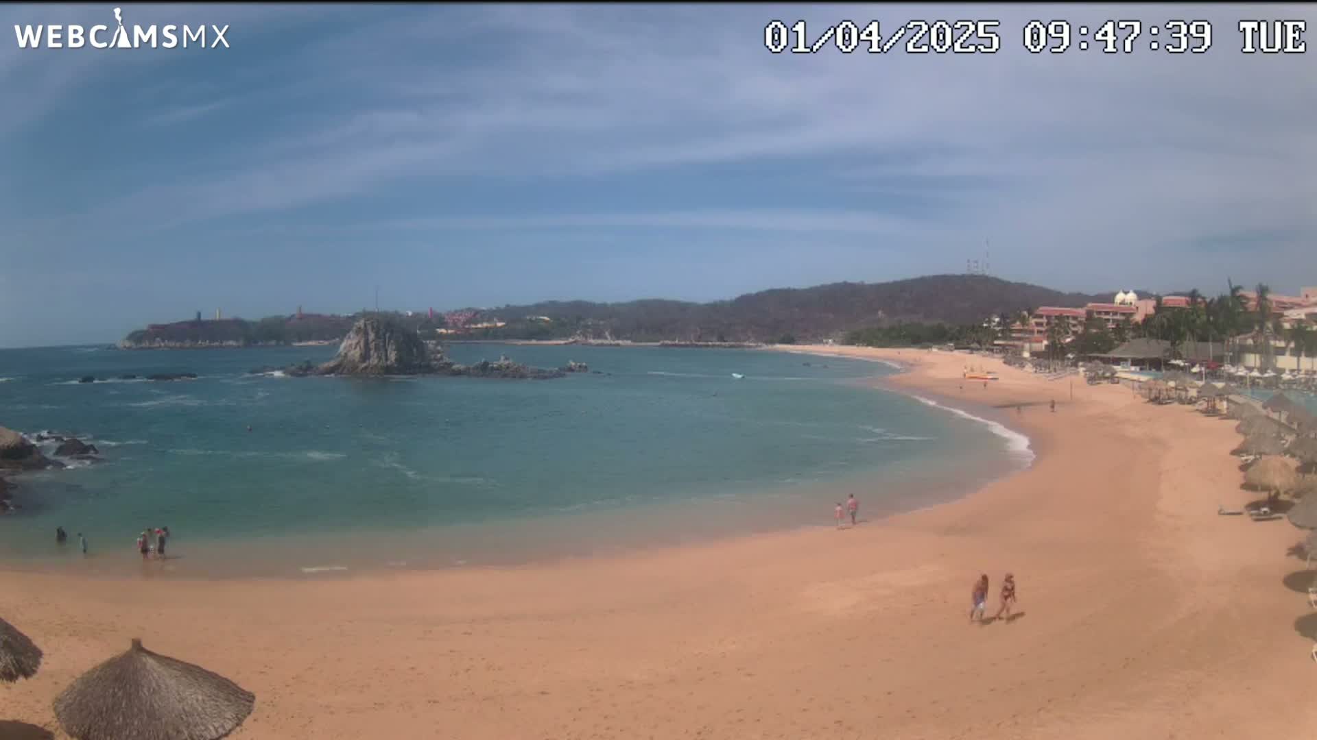 A sandy beach curves around a calm, clear bay under a mostly sunny sky, with a few people enjoying the water and shore.