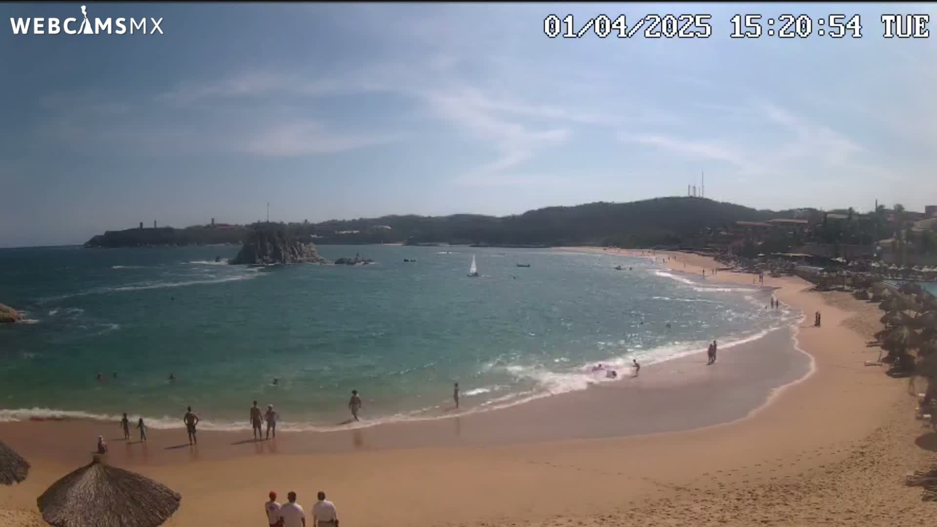 A sandy beach with calm, turquoise water and a few people swimming and sunbathing under a mostly sunny sky.