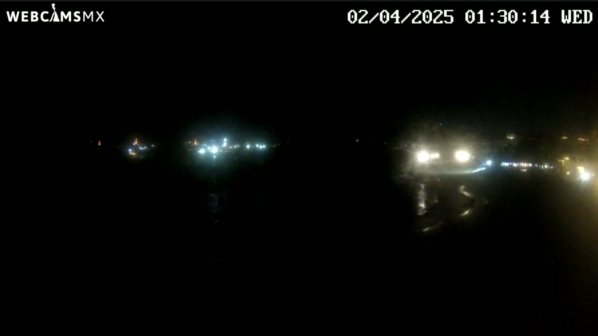A nighttime, low-light view shows a dark body of water reflecting the lights of vehicles and buildings in the distance.