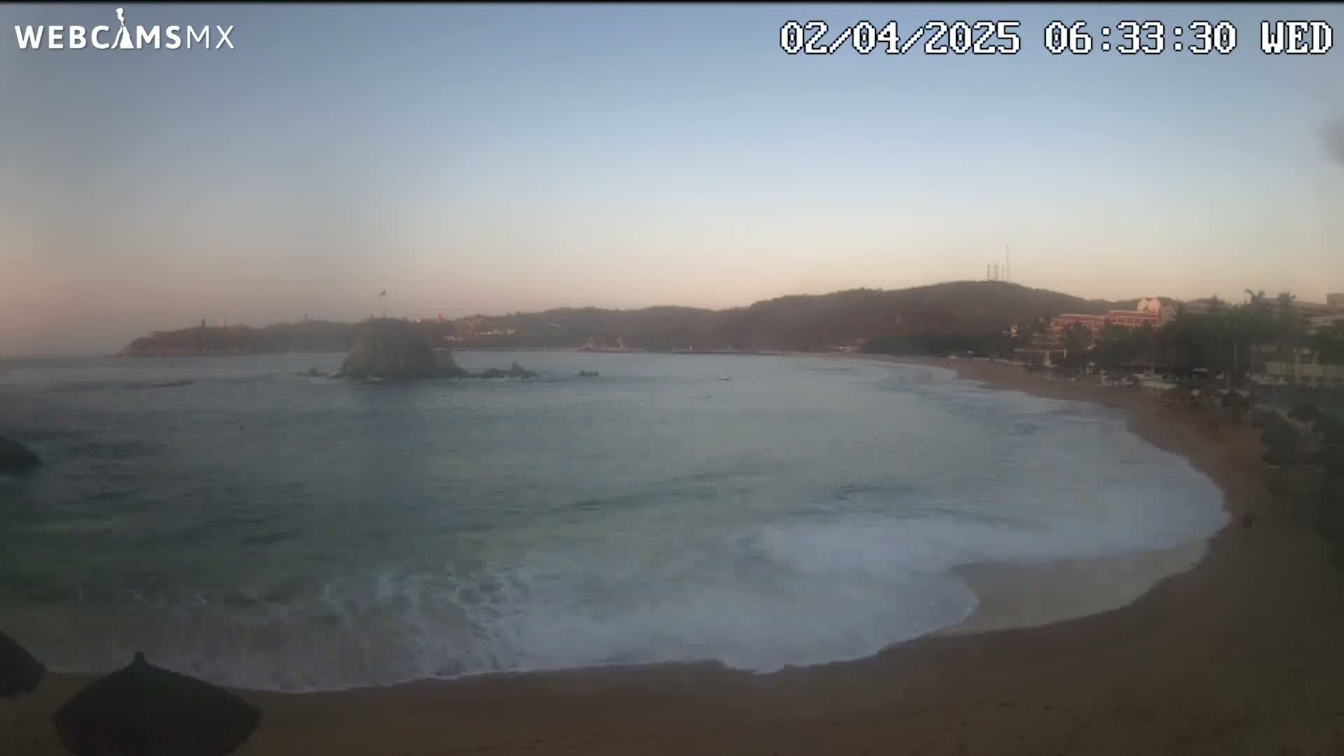 A sandy beach with gentle waves laps the shore under a hazy, dawn sky, with a rocky island and distant hills visible in the background.