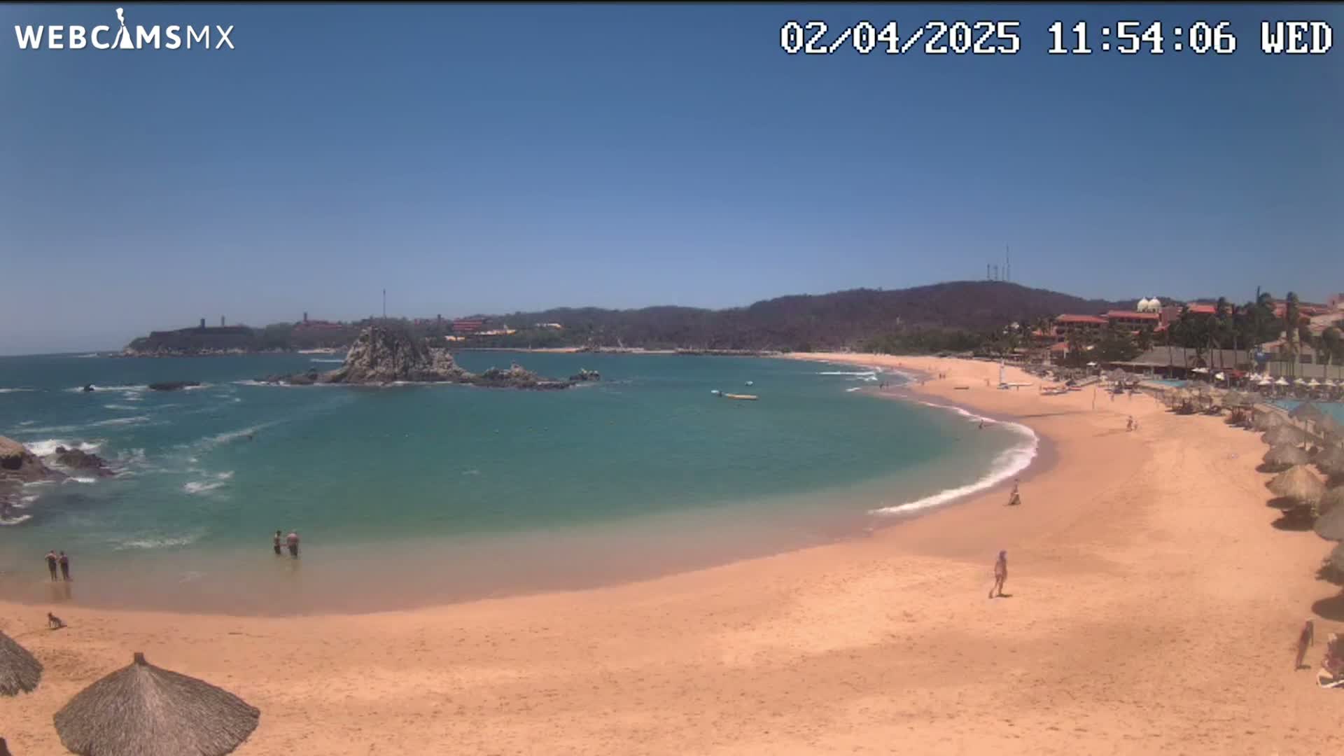 A sandy beach curves around a calm, teal bay under a clear blue sky, with a few people visible on the beach and in the water.