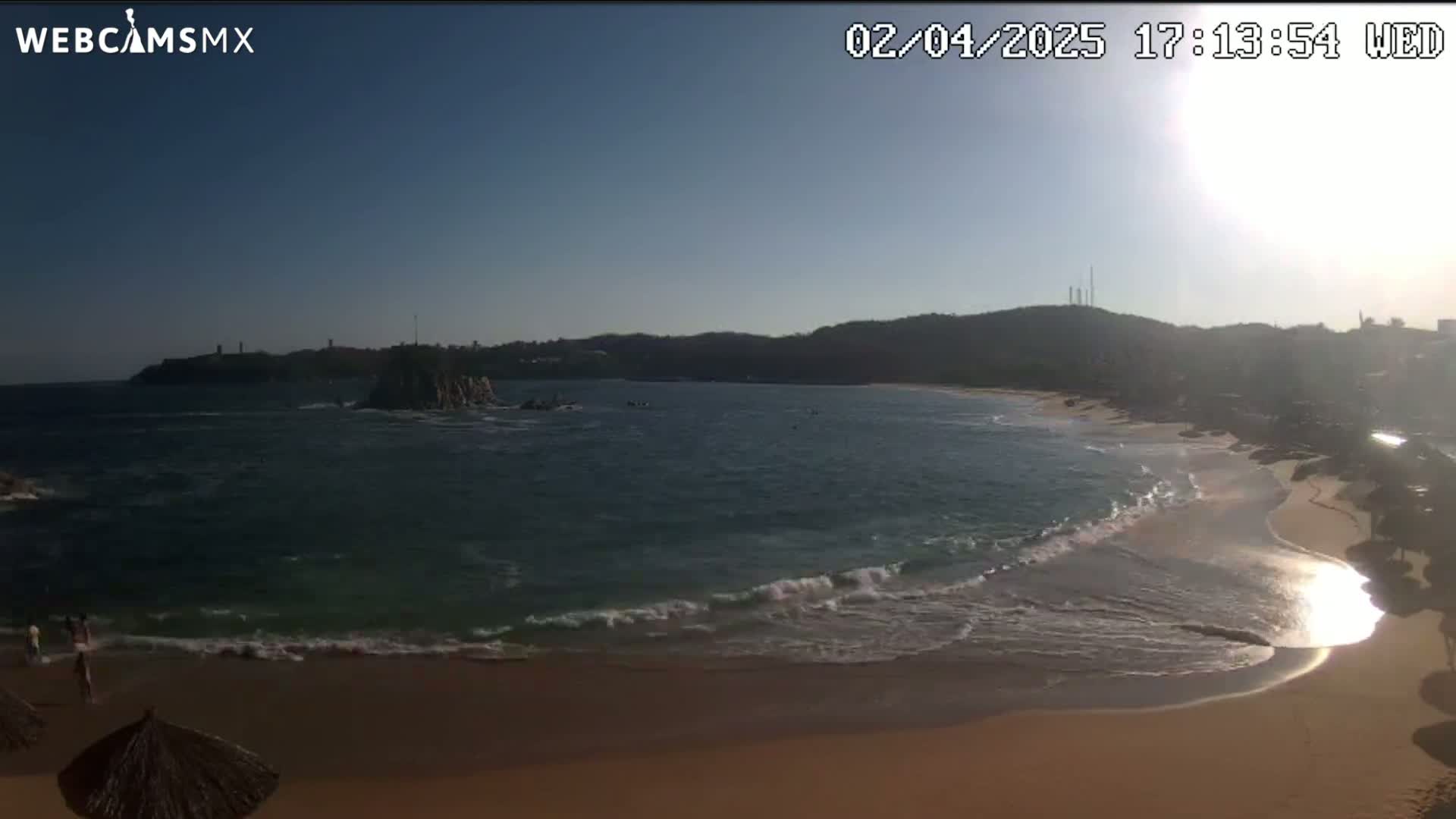 A sandy beach with gentle waves laps the shore under a sunny sky, near a rocky outcropping and a distant hillside.