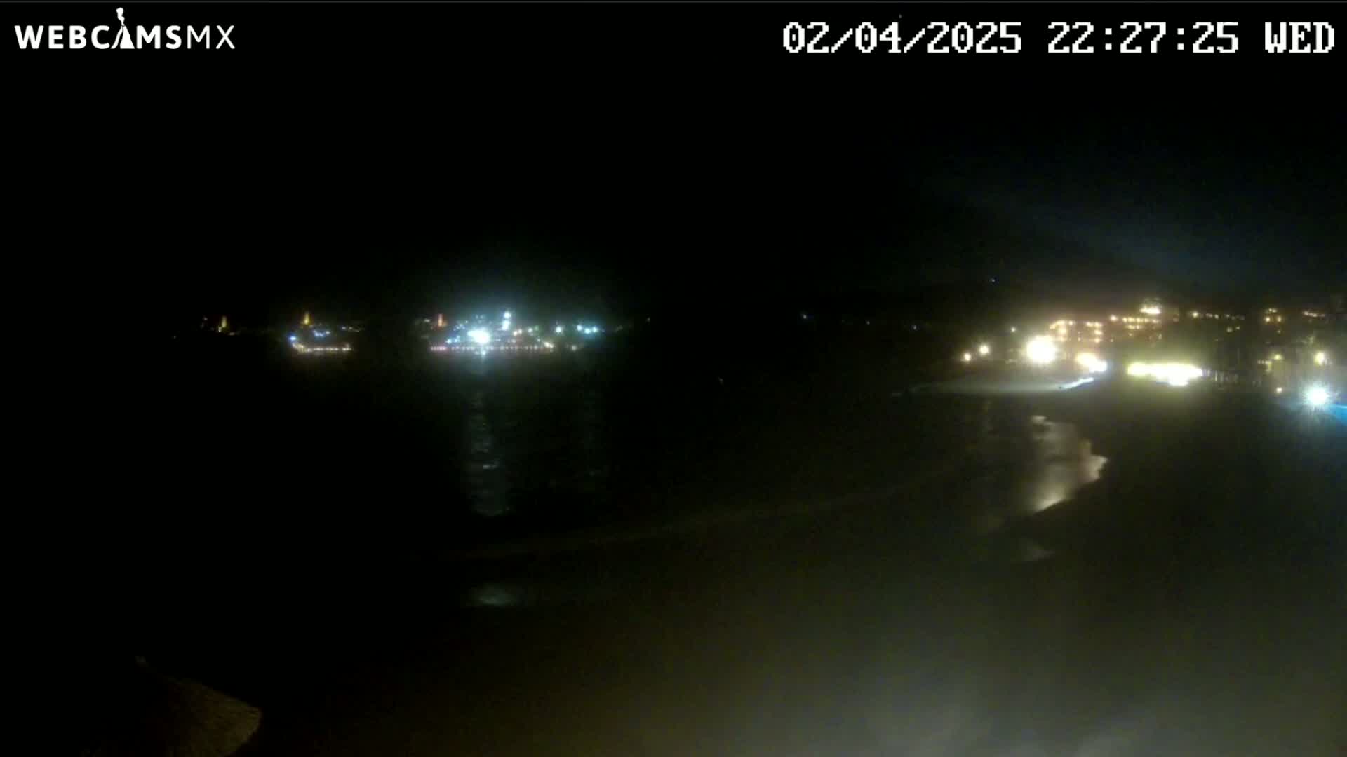 A nighttime long shot of a dark beach and ocean with dimly lit buildings and lights in the distance.