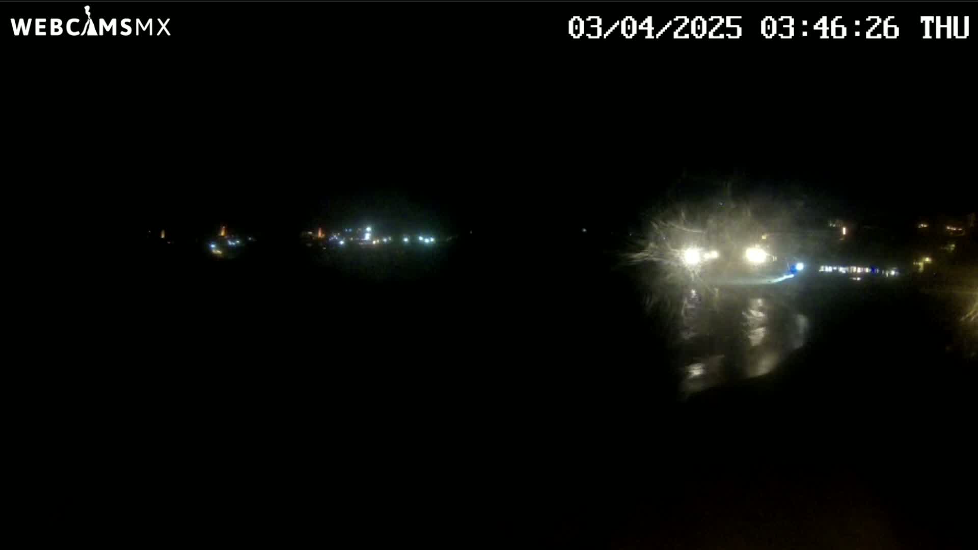 A nighttime view of a wet road with vehicle headlights visible in the foreground, and distant lights indicating a town or city in the background under a dark, possibly stormy sky.