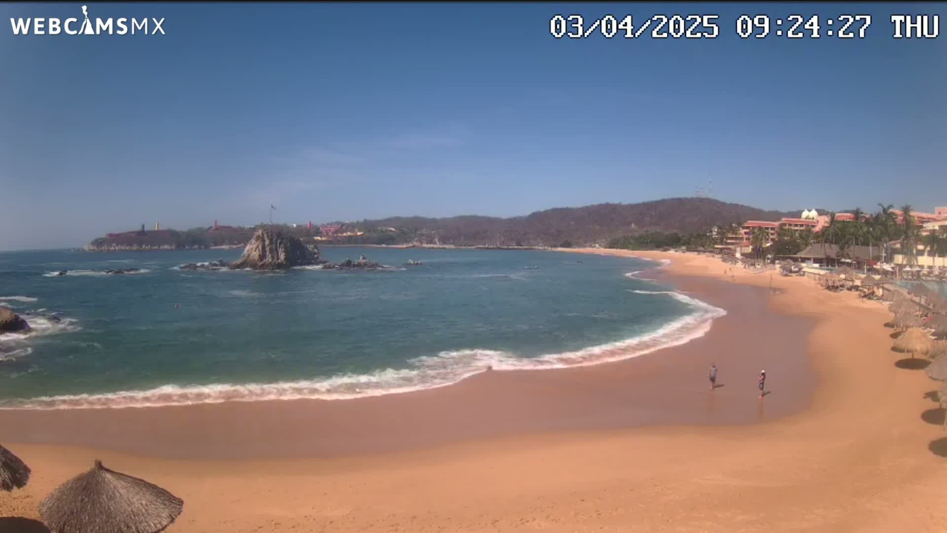 A sandy beach with calm waves laps the shore under a clear blue sky, with a rocky island and distant buildings visible across the bay.
