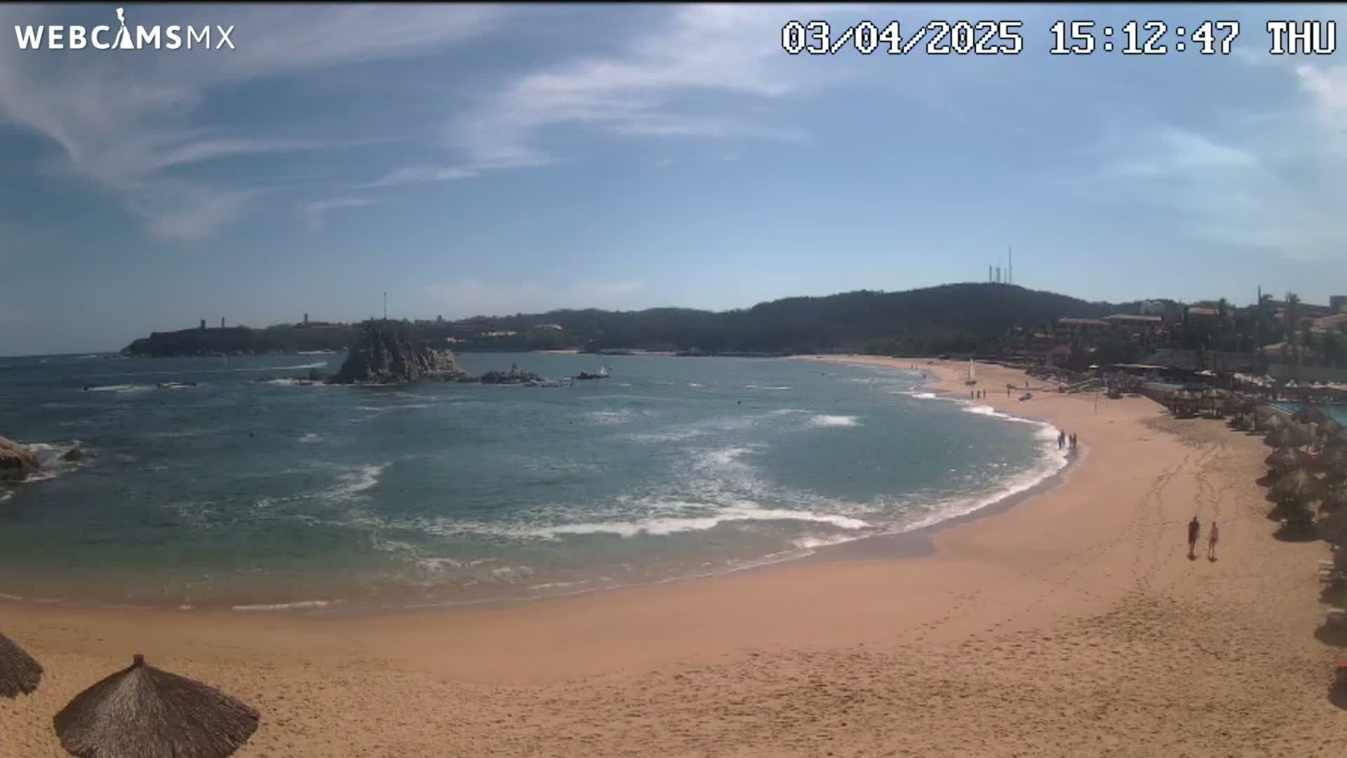 A sandy beach curves around a calm bay with a rocky outcropping in the water under a mostly sunny sky.