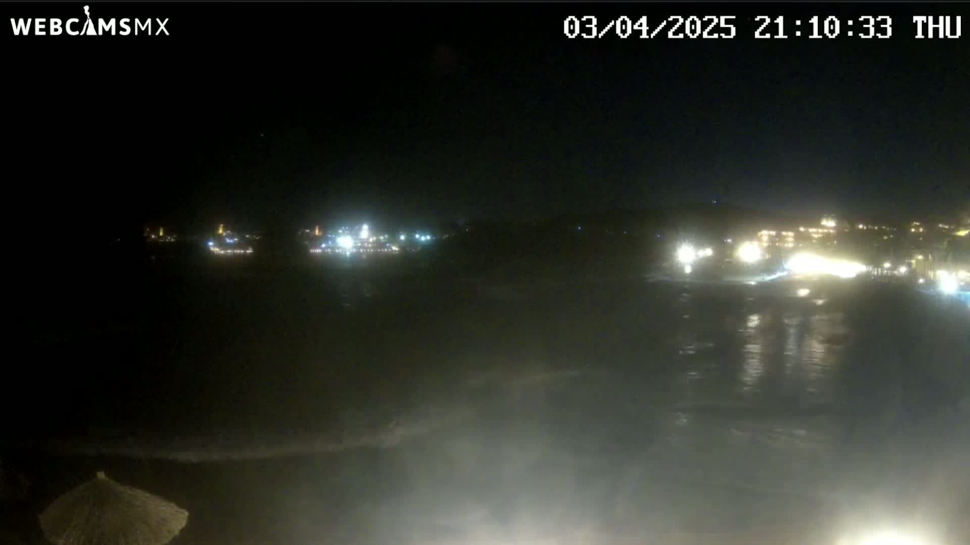 A nighttime view of a dark ocean with waves lapping at the shore, illuminated by lights from buildings on a distant coastline under a clear sky.