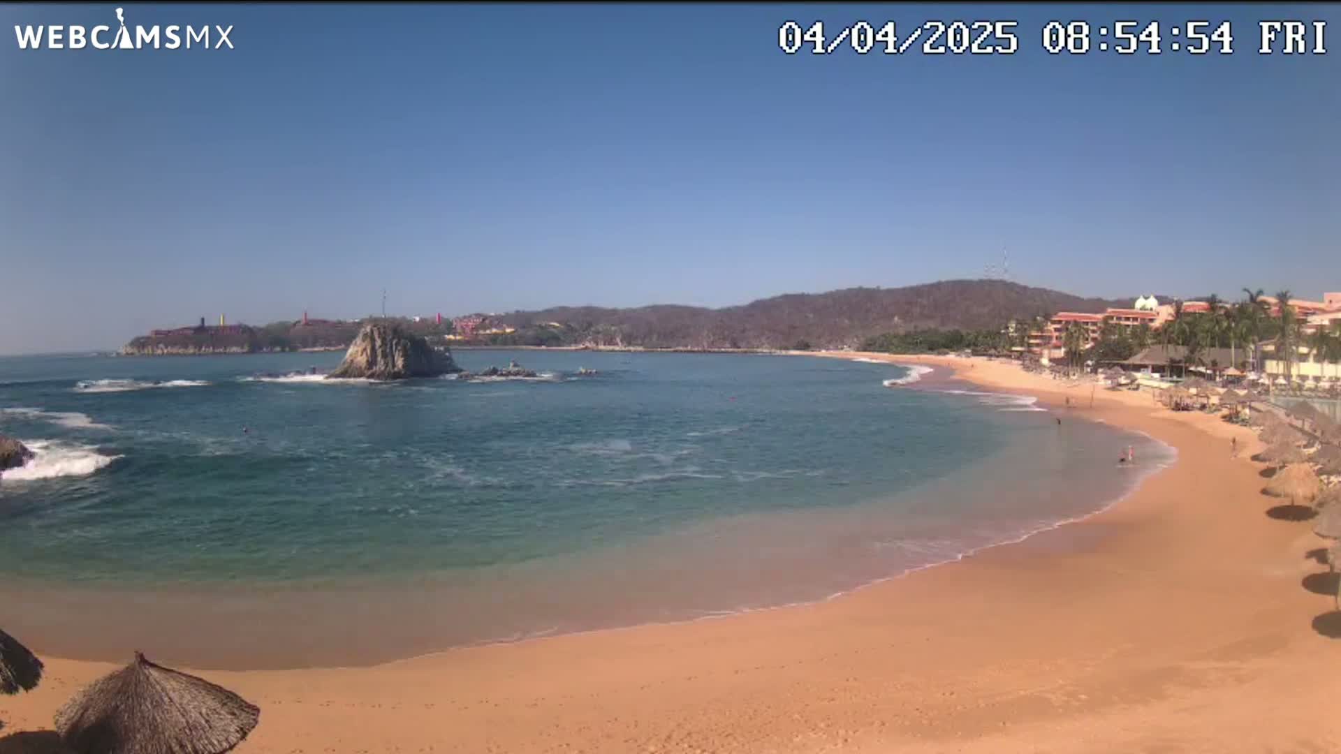 A sandy beach curves around a calm, clear bay under a bright, sunny sky, with a rocky islet and distant buildings visible.