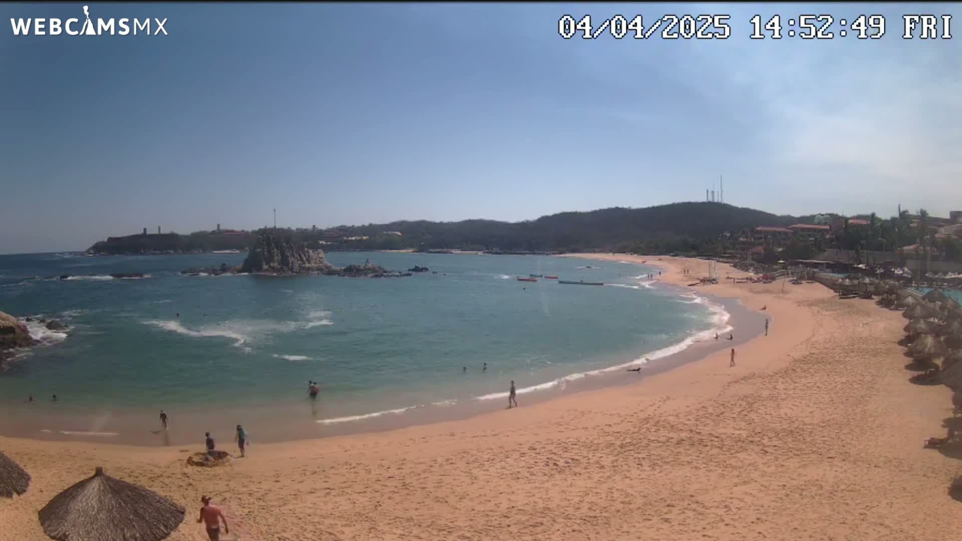 A sandy beach curves around a calm, teal ocean under a clear blue sky, with a few people swimming and sunbathing.