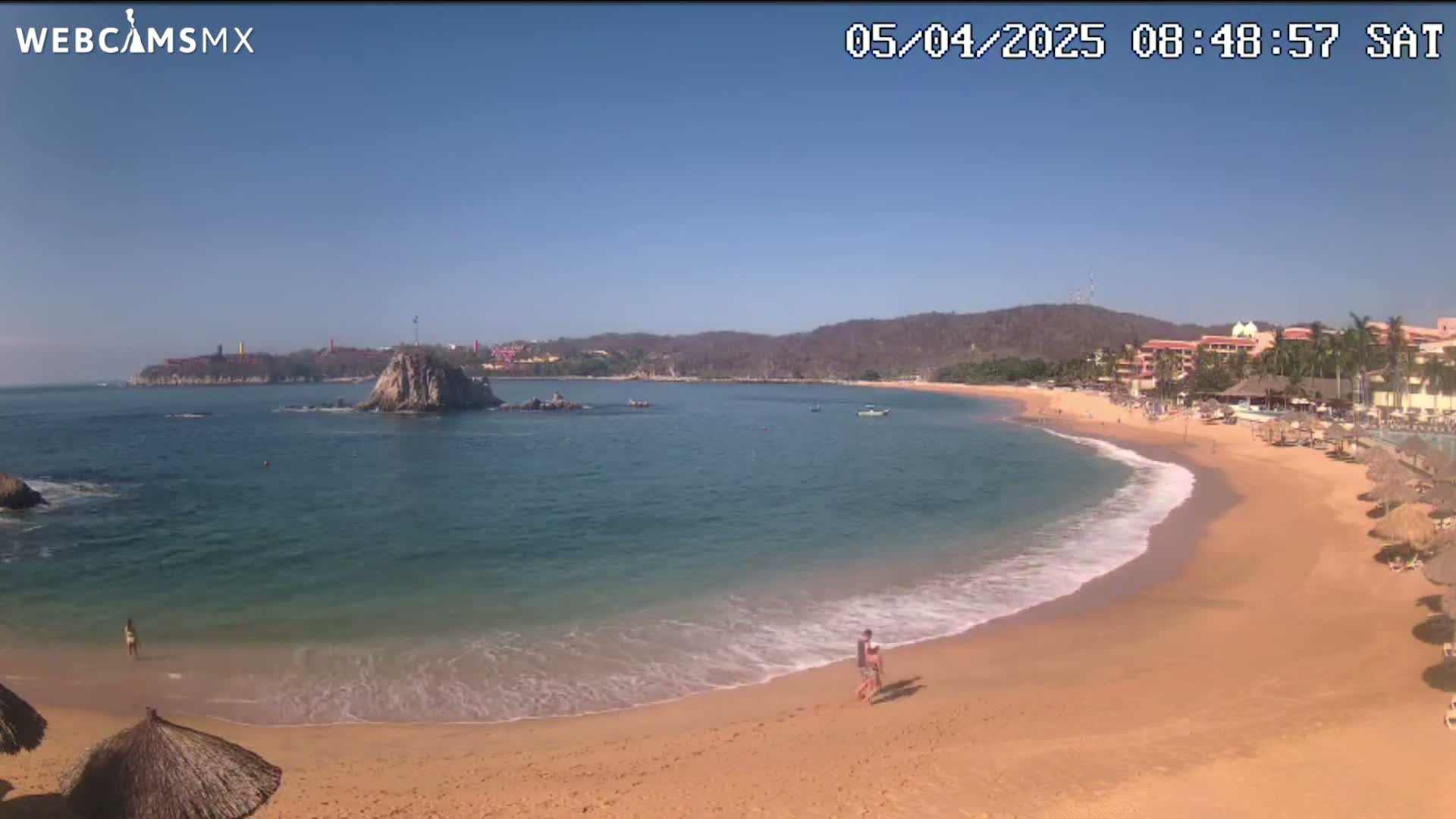 A sandy beach curves around a calm, turquoise bay under a clear blue sky, with a few people walking along the shore and some buildings visible in the distance.