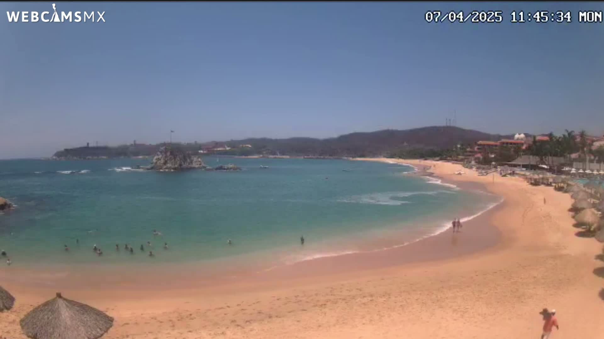 A sandy beach with calm, clear turquoise water laps the shore under a clear blue sky, with a few people swimming and walking along the beach.