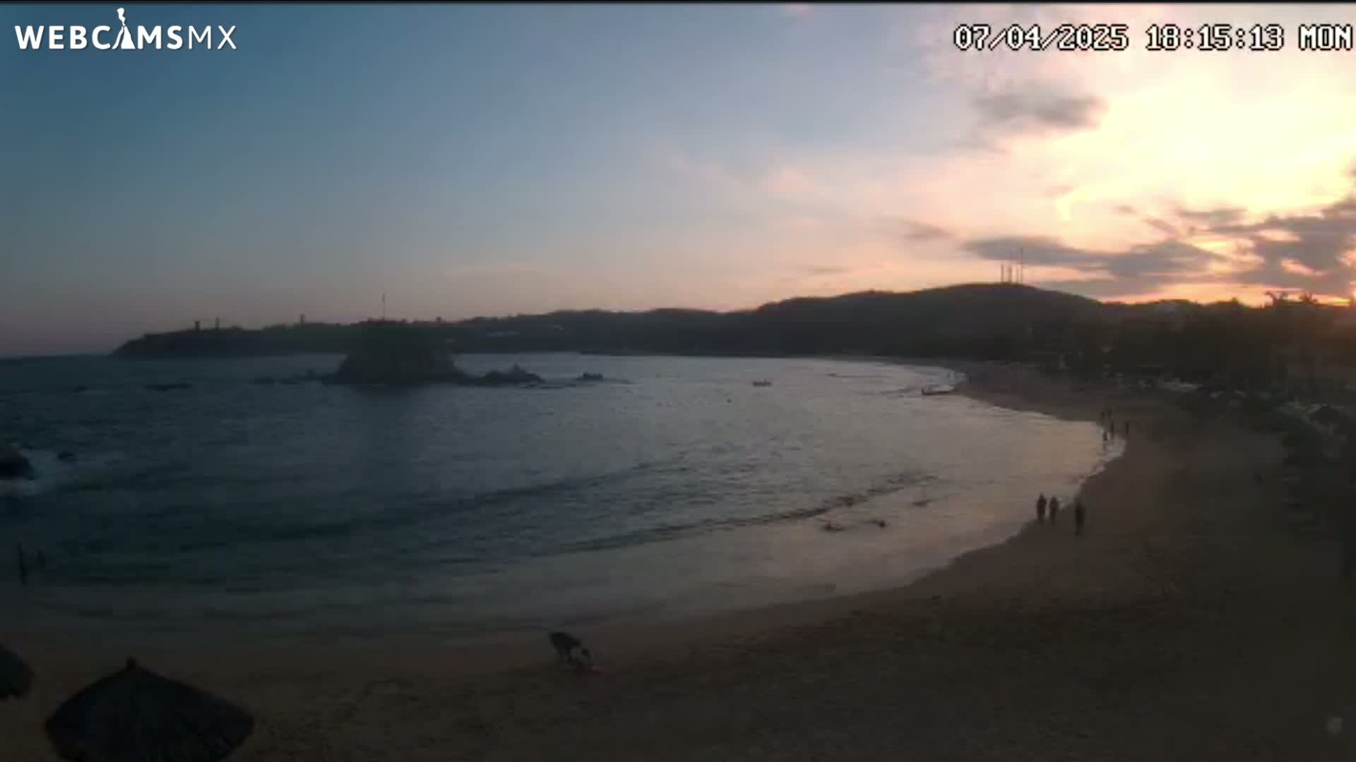 A sandy beach at sunset with calm ocean waves and a few people walking along the shore.