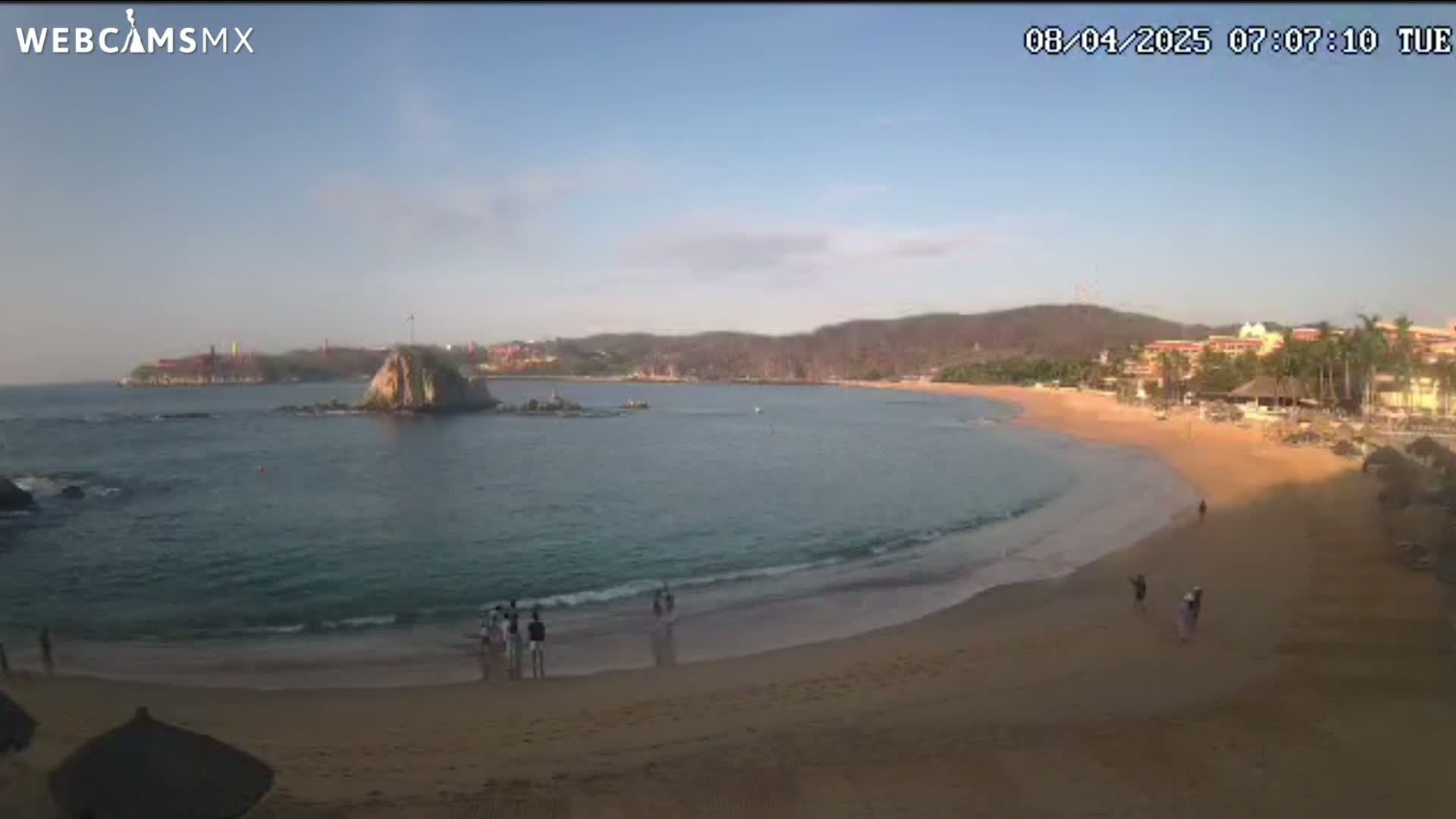 A sandy beach curves around a calm bay, with a rocky islet in the water and distant hills under a clear sky.