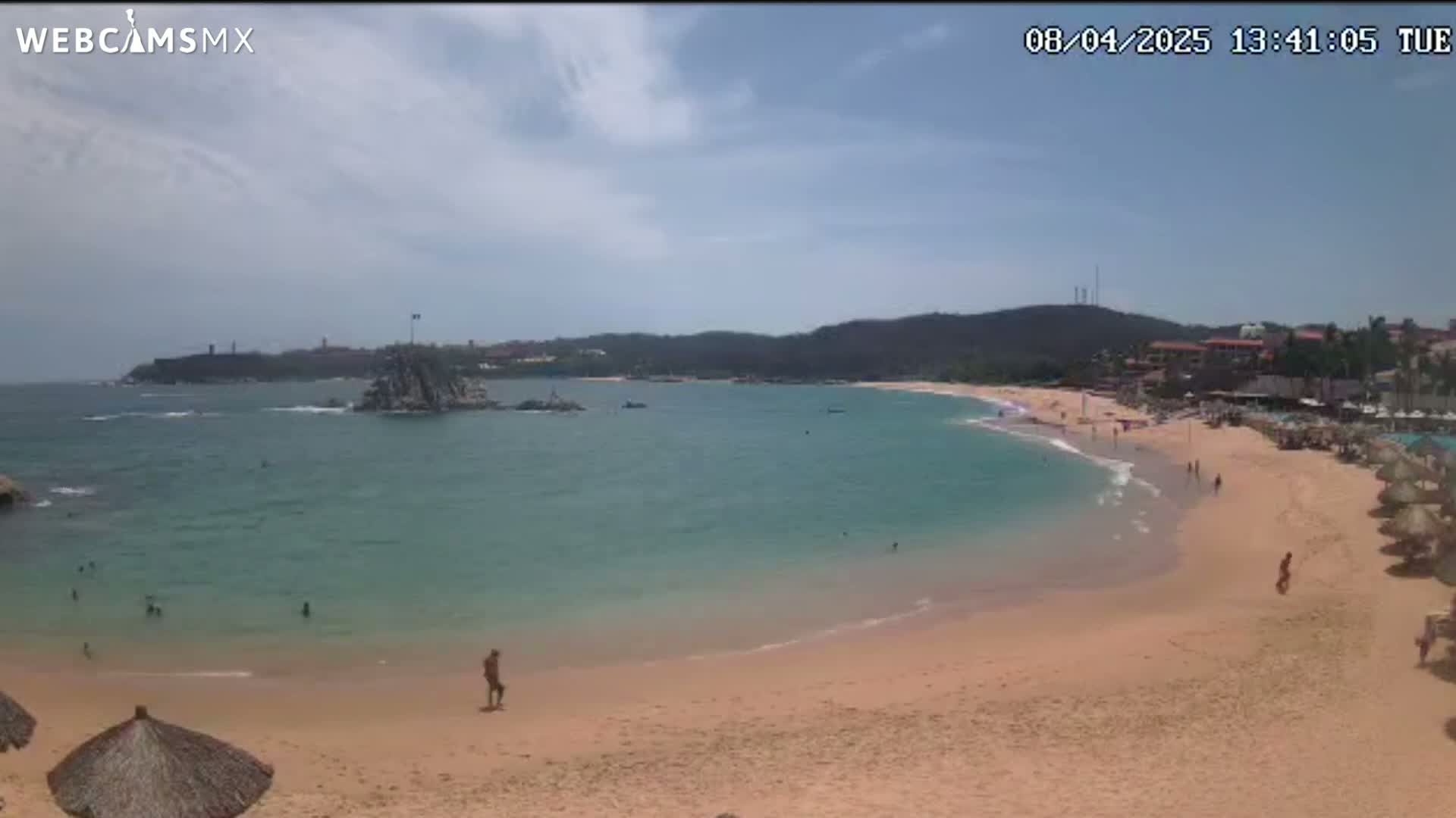 A partly sunny day shows a secluded, sandy beach curving around a calm, turquoise bay with a rocky islet and distant hills.