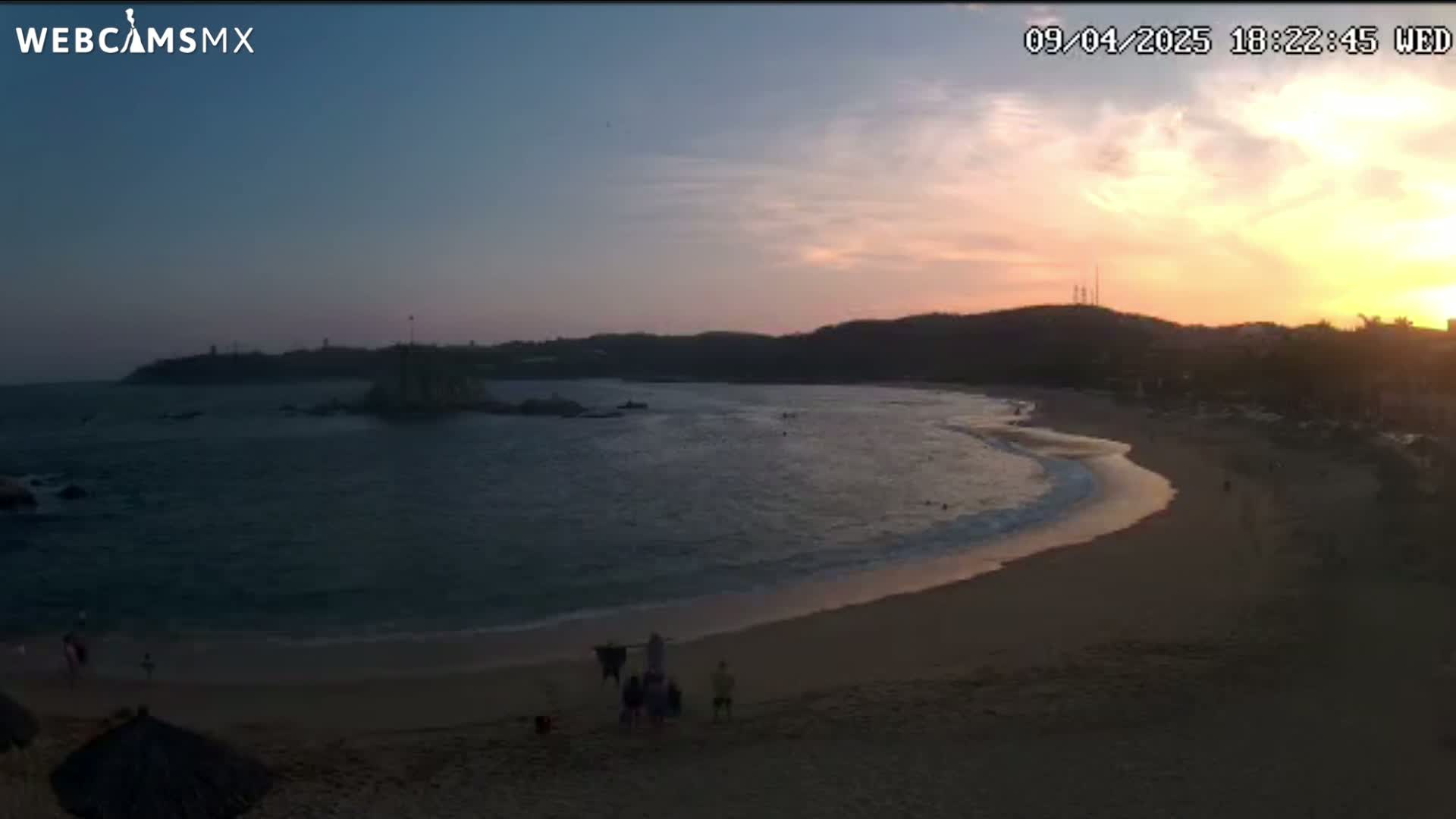 A sandy beach curves into a calm ocean at sunset under a partly cloudy sky.