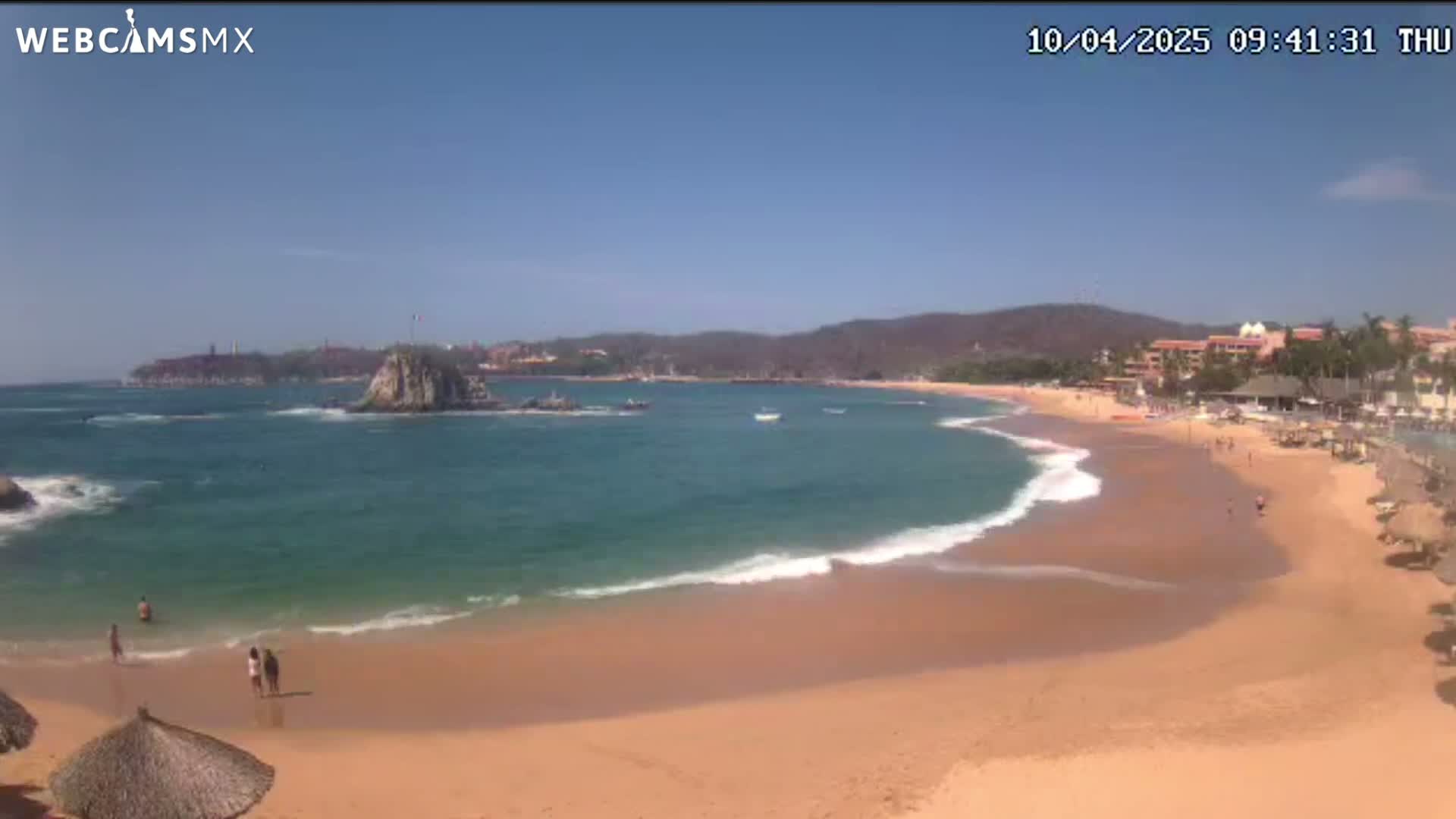 A sandy beach curves around a calm, teal ocean under a clear blue sky, with a rocky outcropping and distant buildings visible.