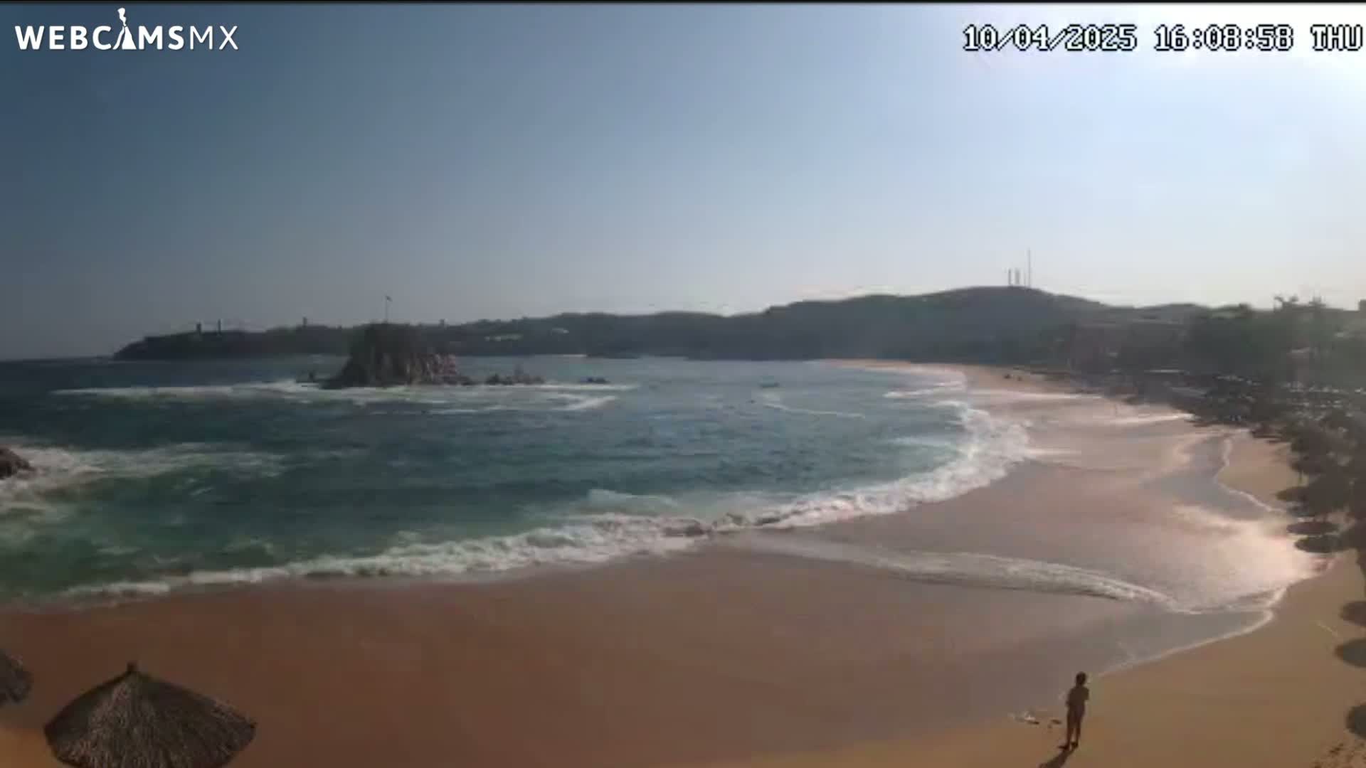A sandy beach with gentle waves laps the shore under a clear, sunny sky, with a rocky outcrop visible in the ocean and buildings in the distance.
