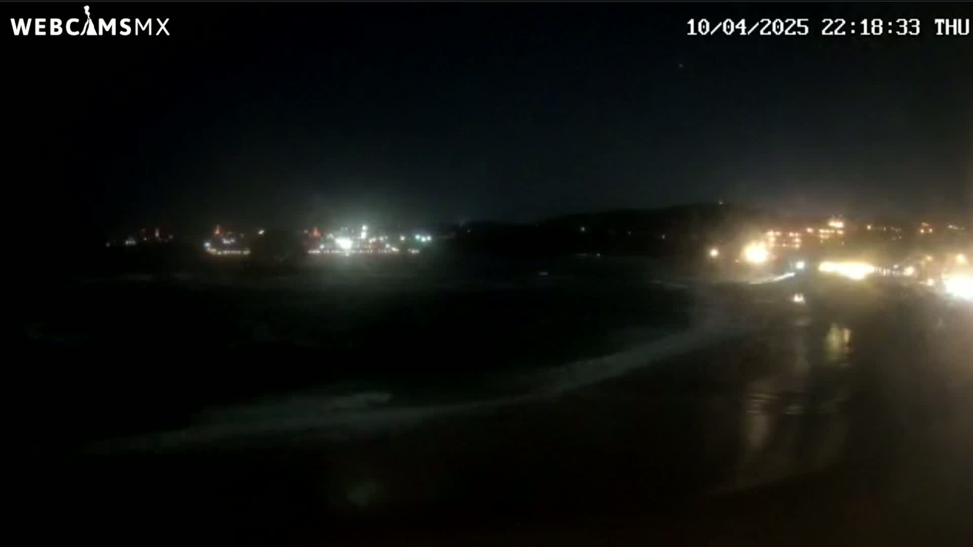 A nighttime view of a dark beach with small waves, illuminated by distant city lights.