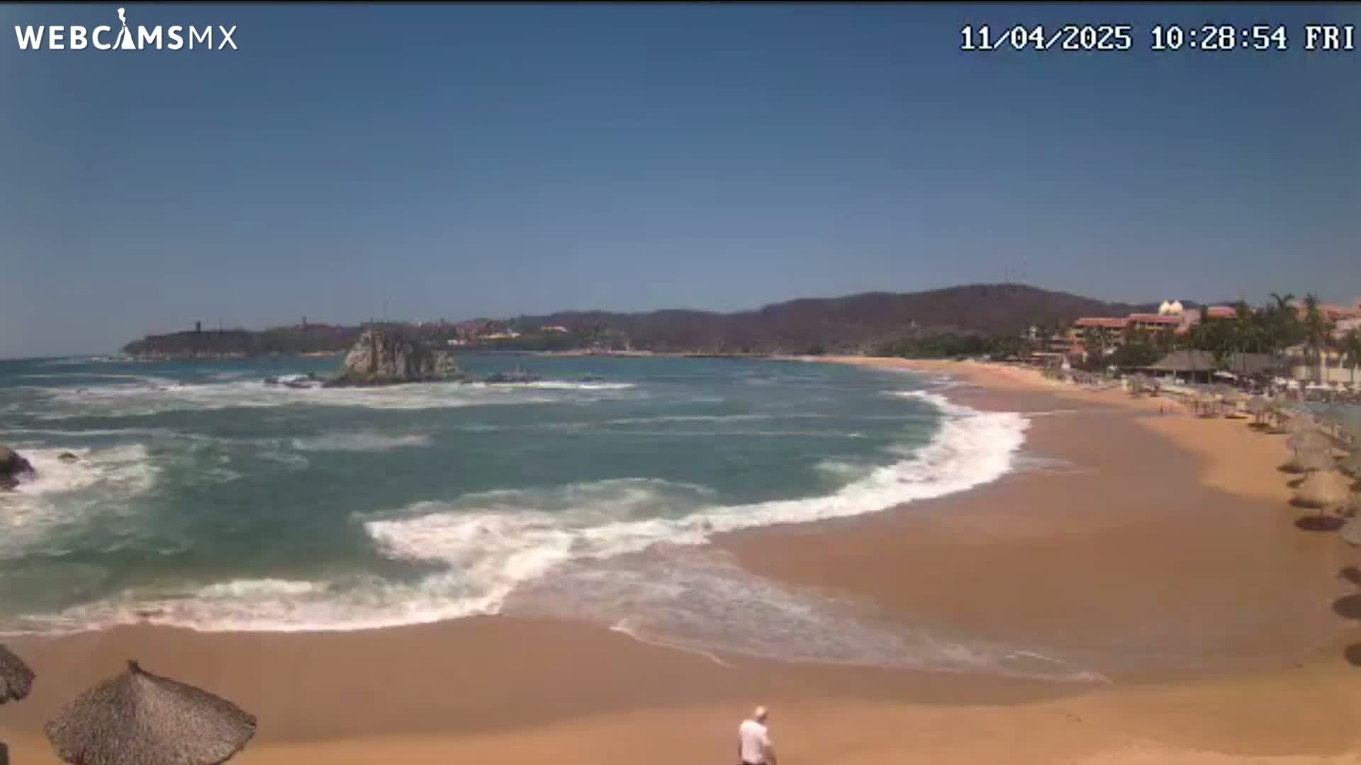 A sandy beach with moderate waves under a clear blue sky stretches out to a rocky coastline and distant hills.