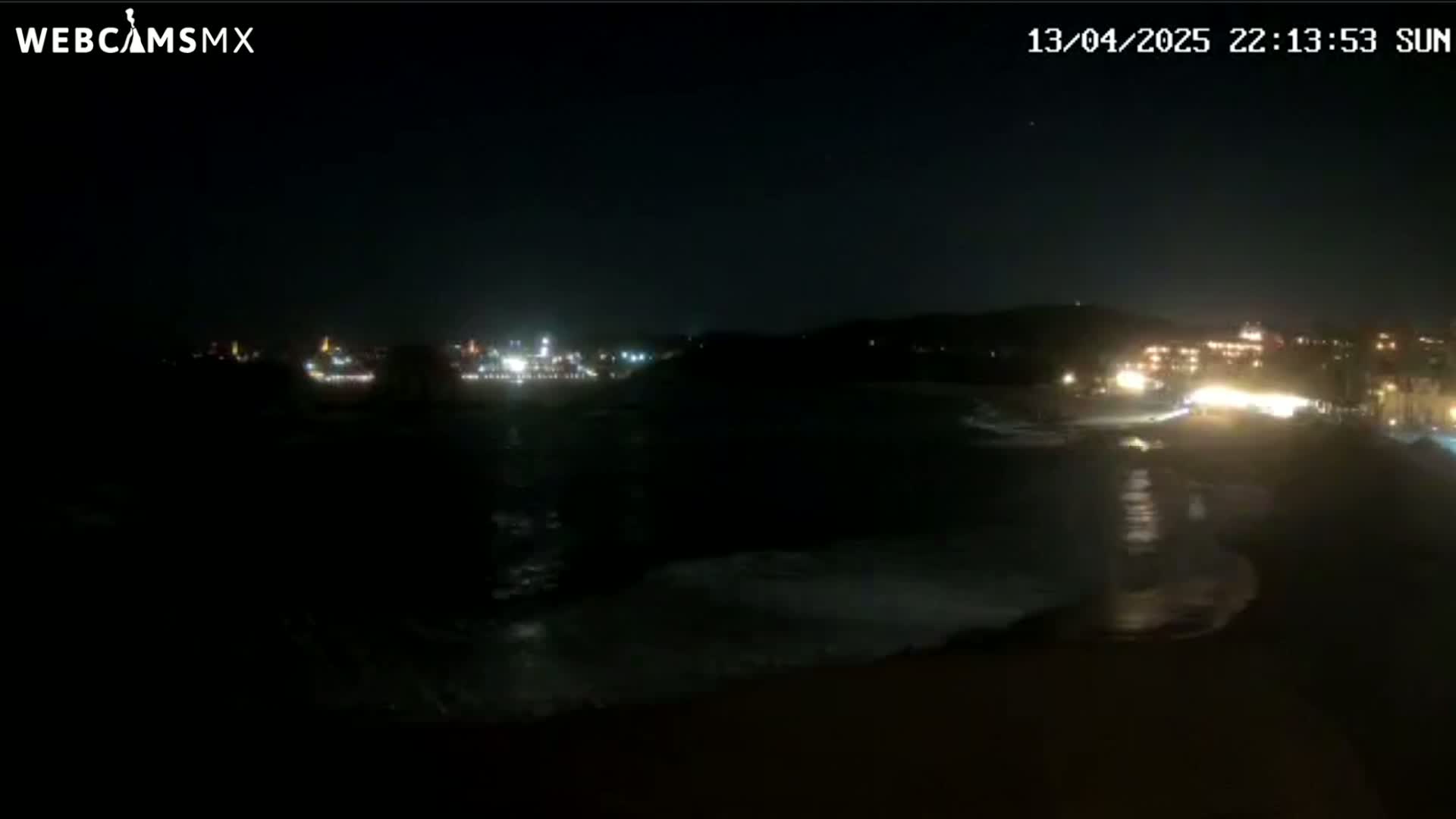 A nighttime, seaside view shows dark water lapping a beach with a faintly lit town in the distance.