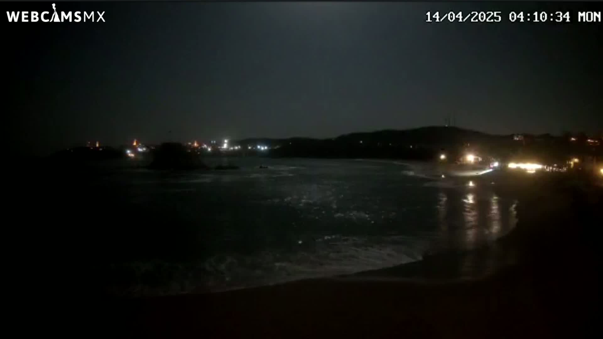 A dark nighttime beach scene shows a moonlit ocean with waves lapping the shore, under a clear sky, with distant lights illuminating a coastline.