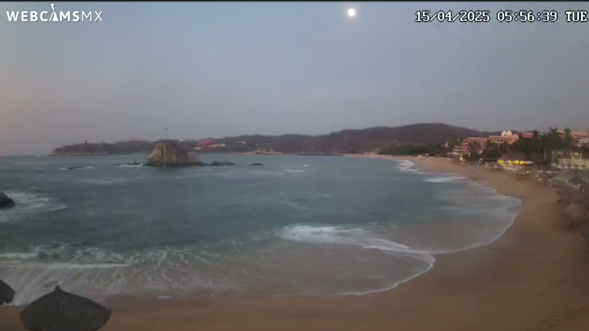 A sandy beach with gentle waves rolls onto the shore, under a mostly cloudy sky, with a rocky island visible in the calm bay.
