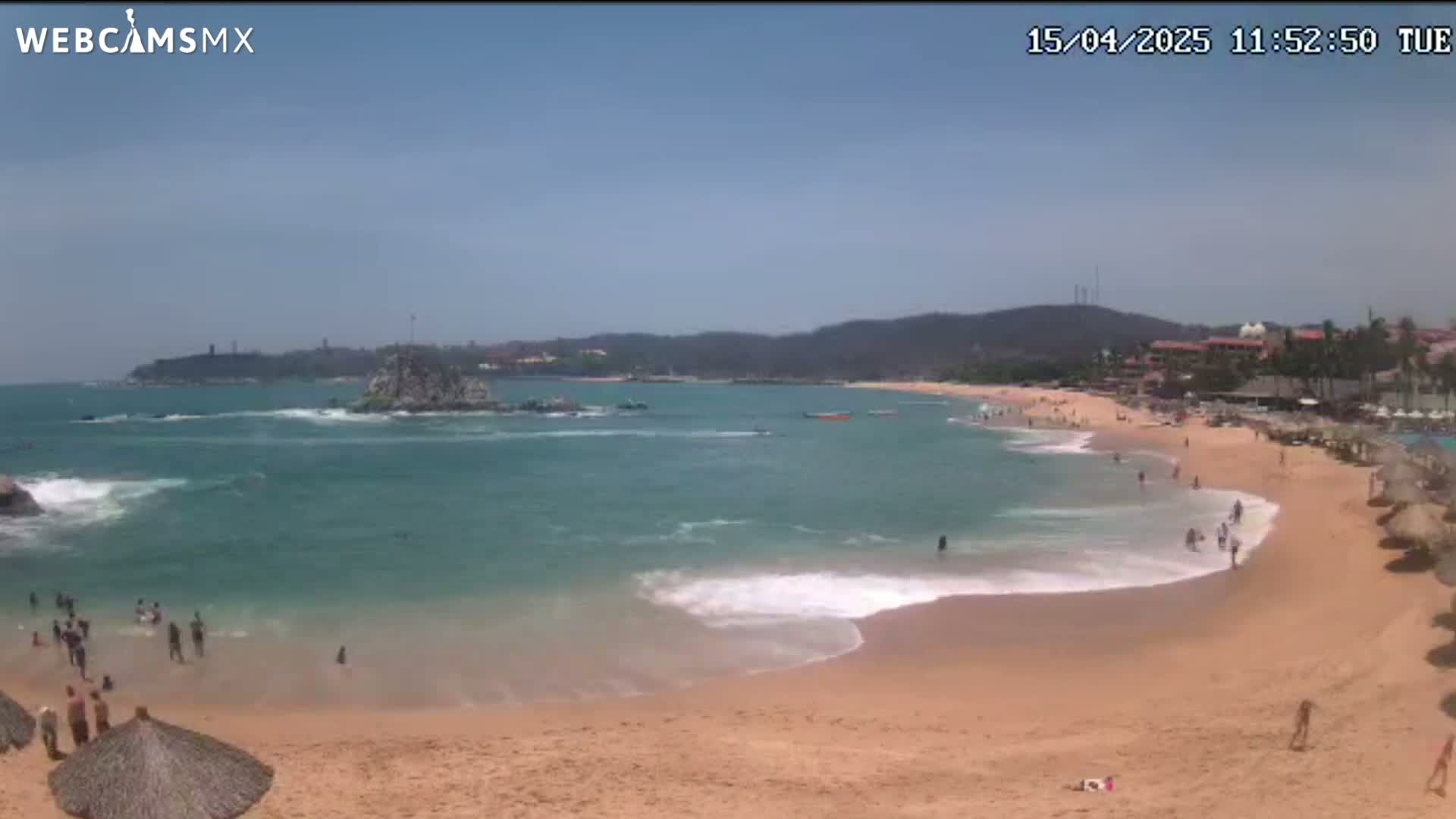 A sandy beach with calm, turquoise water laps gently at the shore under a clear blue sky, with people swimming and relaxing on the sand and a rocky outcropping in the ocean.