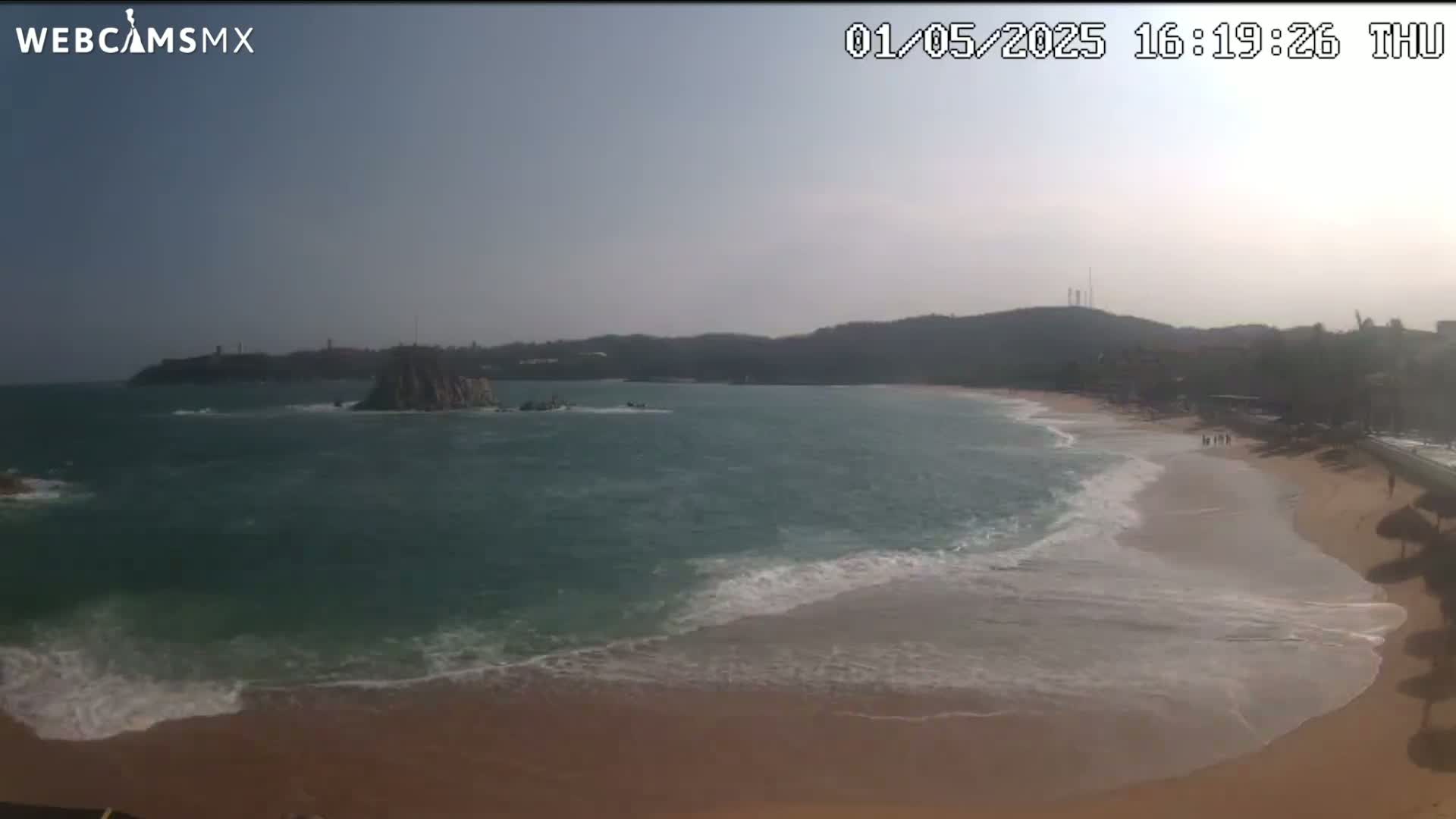 A sandy beach with gentle waves laps the shore under a mostly clear sky, with a rocky island visible in the ocean.