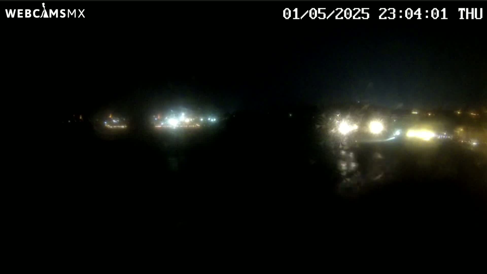 A nighttime view of a road with vehicle headlights visible in the distance, under dark and possibly foggy or rainy conditions.