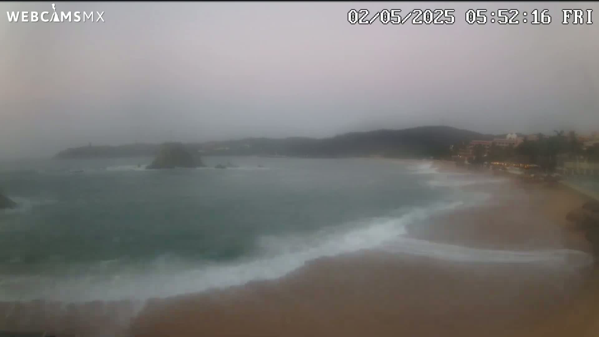 A beach with stormy, overcast weather shows waves crashing on the shore near buildings and hills in the distance.