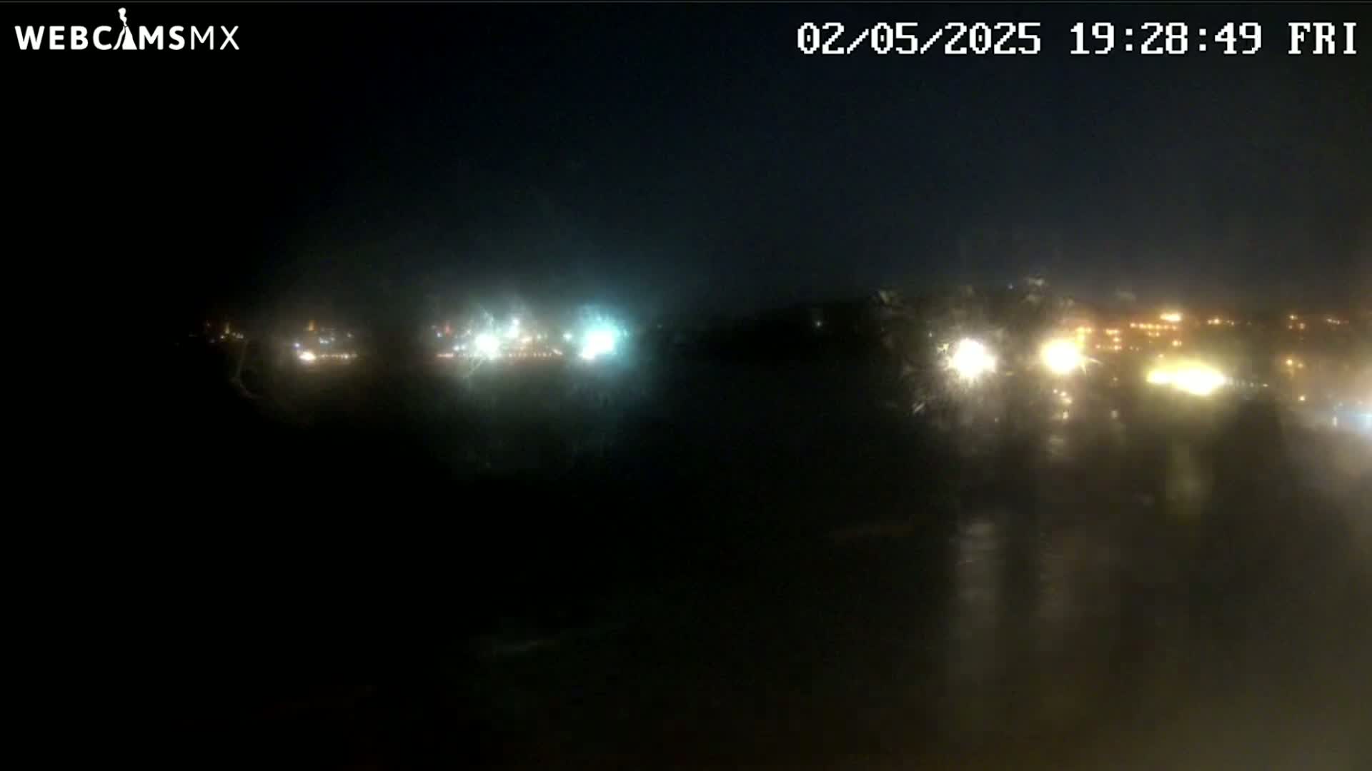A nighttime, rain-streaked view of distant city lights across a dark body of water.
