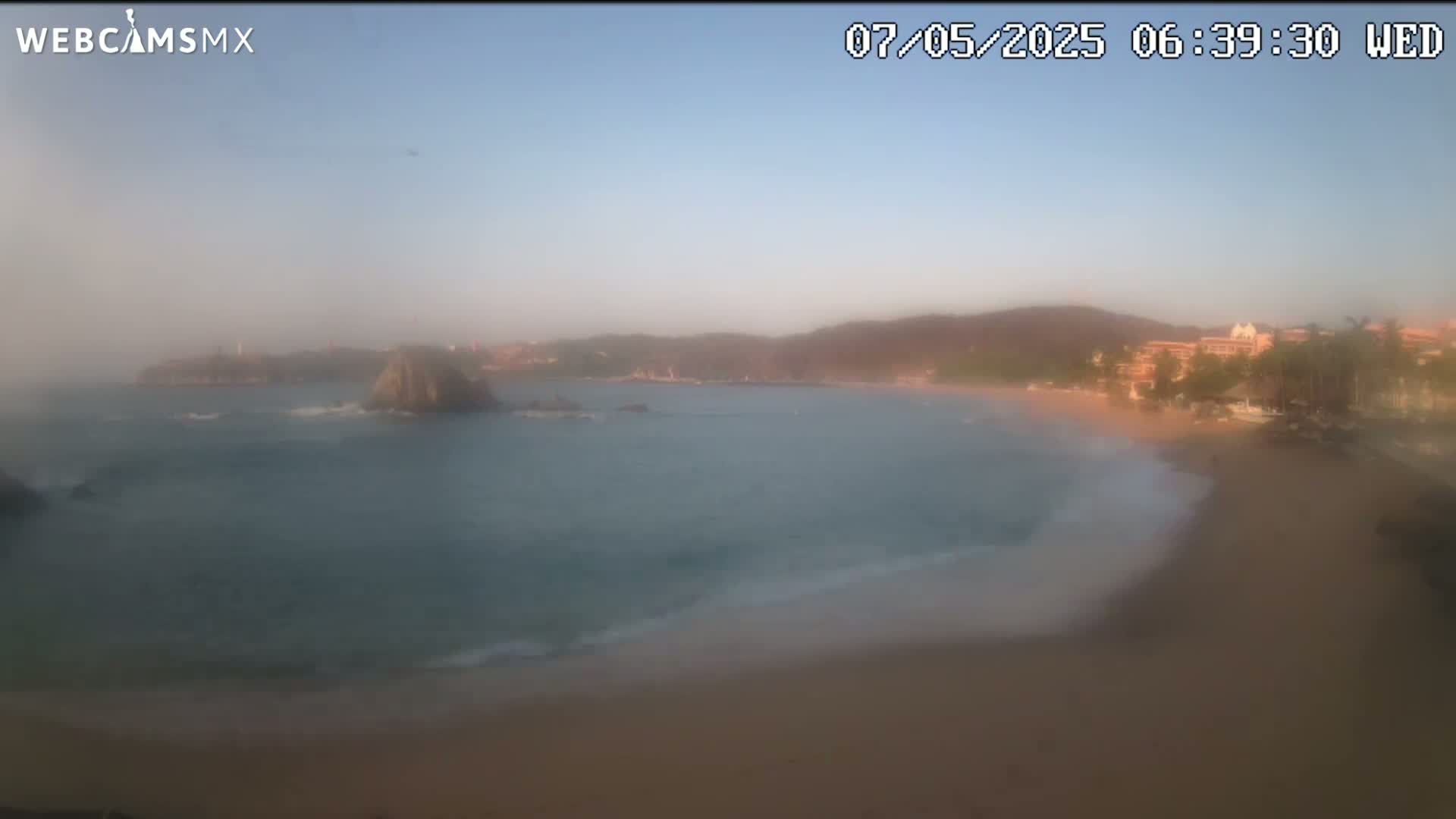 A hazy sunrise illuminates a sandy beach and calm ocean, with a rocky outcrop and distant hills visible in the background.