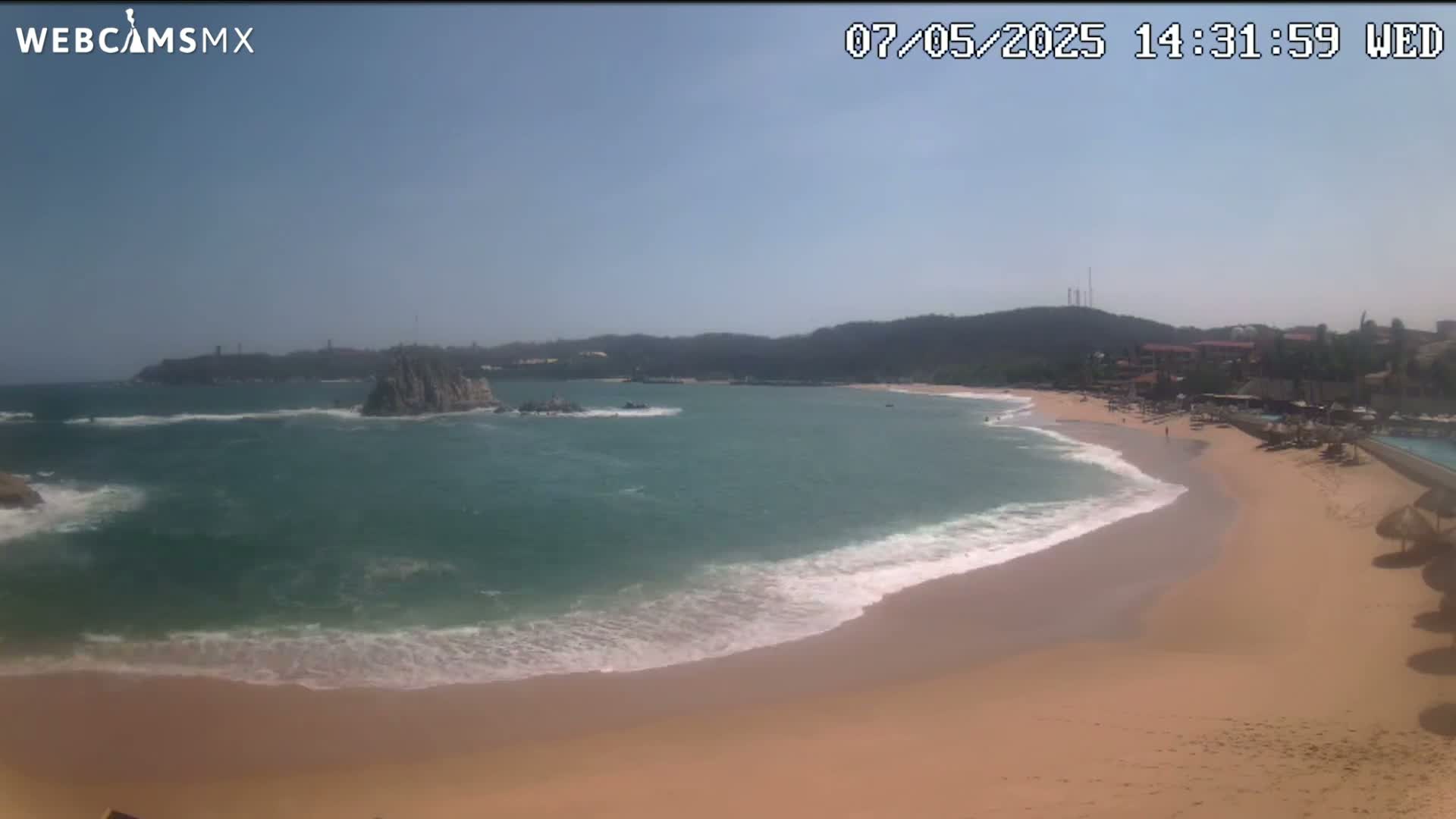 A sandy beach with gentle waves laps at the shore under a clear, sunny sky, backed by a calm bay and distant hills.
