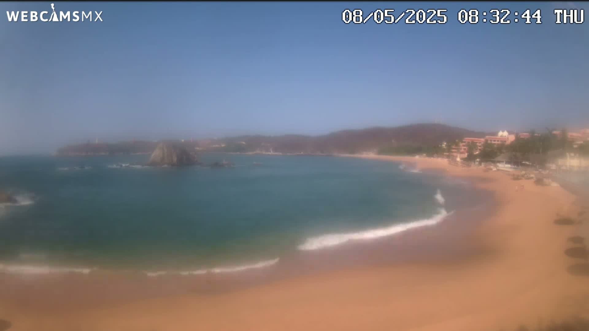 A sandy beach curves around a calm bay with small waves lapping the shore, under a clear blue sky.