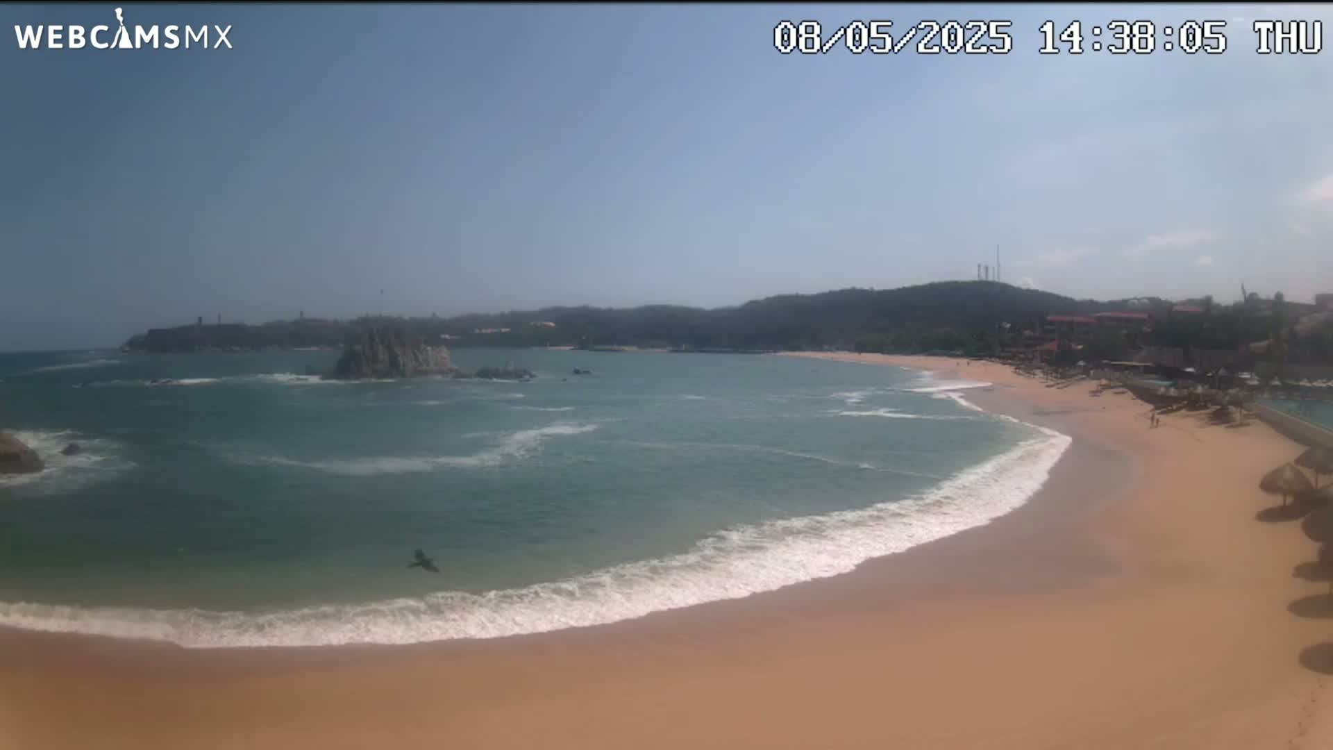 A sandy beach curves around a calm, teal ocean with small waves lapping the shore under a clear, sunny sky.