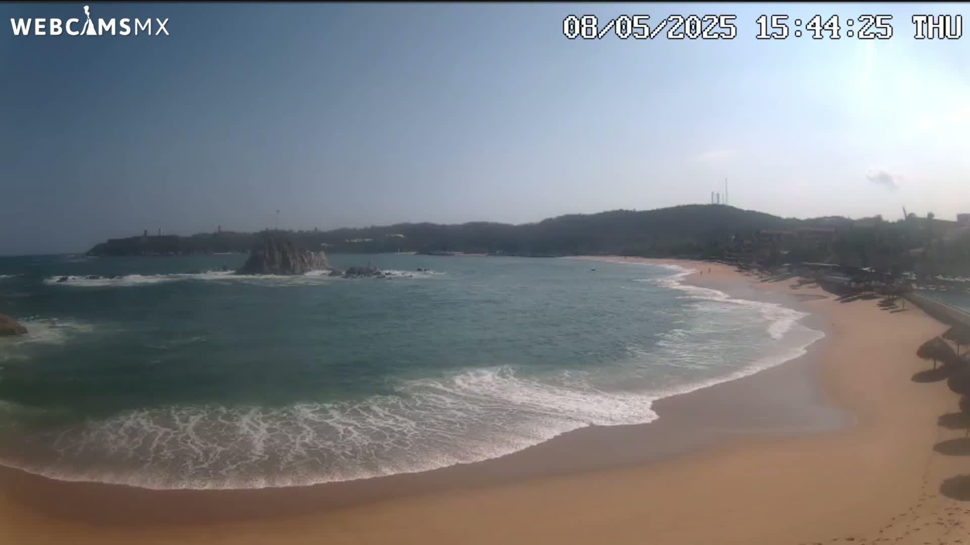 A sandy beach with gentle waves laps the shore under a clear, sunny sky, with a rocky outcrop in the calm bay beyond.