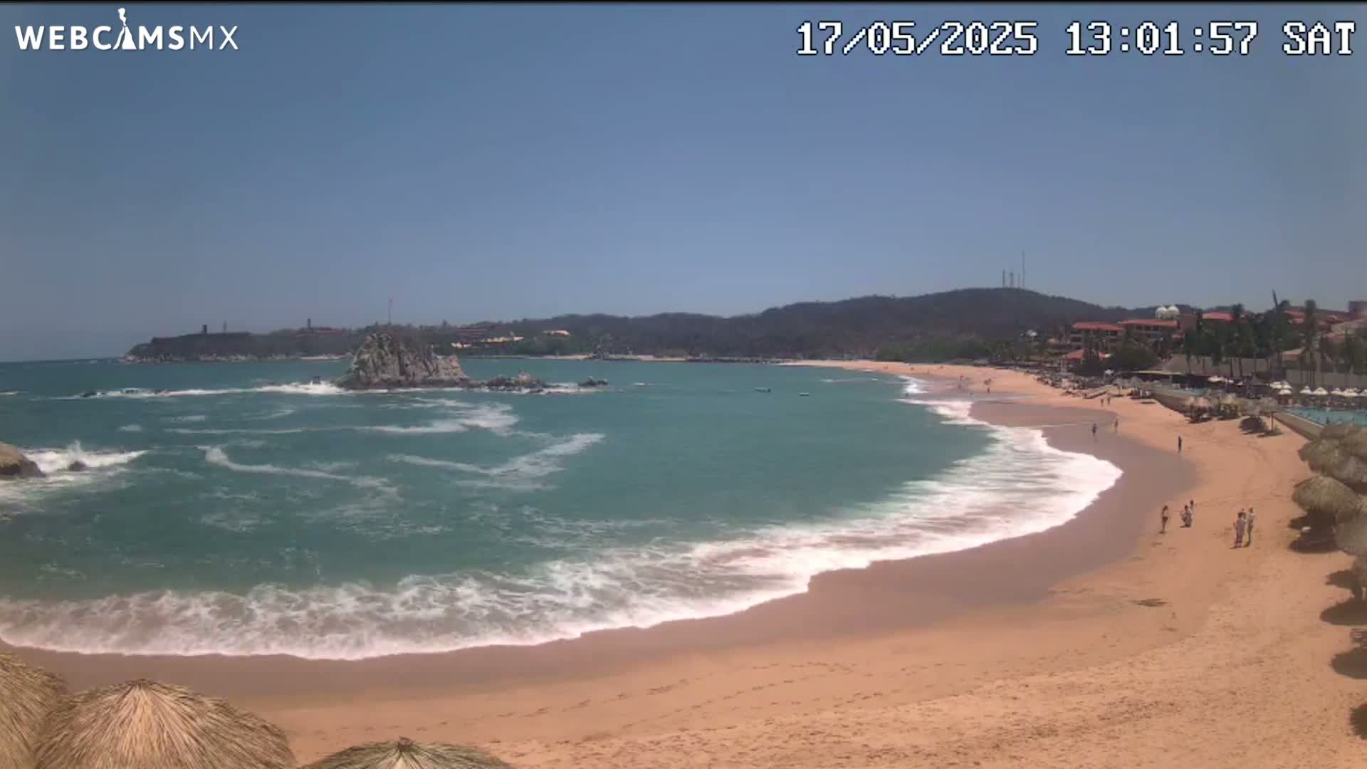 A sandy beach with gentle waves laps the shore under a clear, sunny sky, with a rocky outcrop in the ocean and a resort visible in the background.