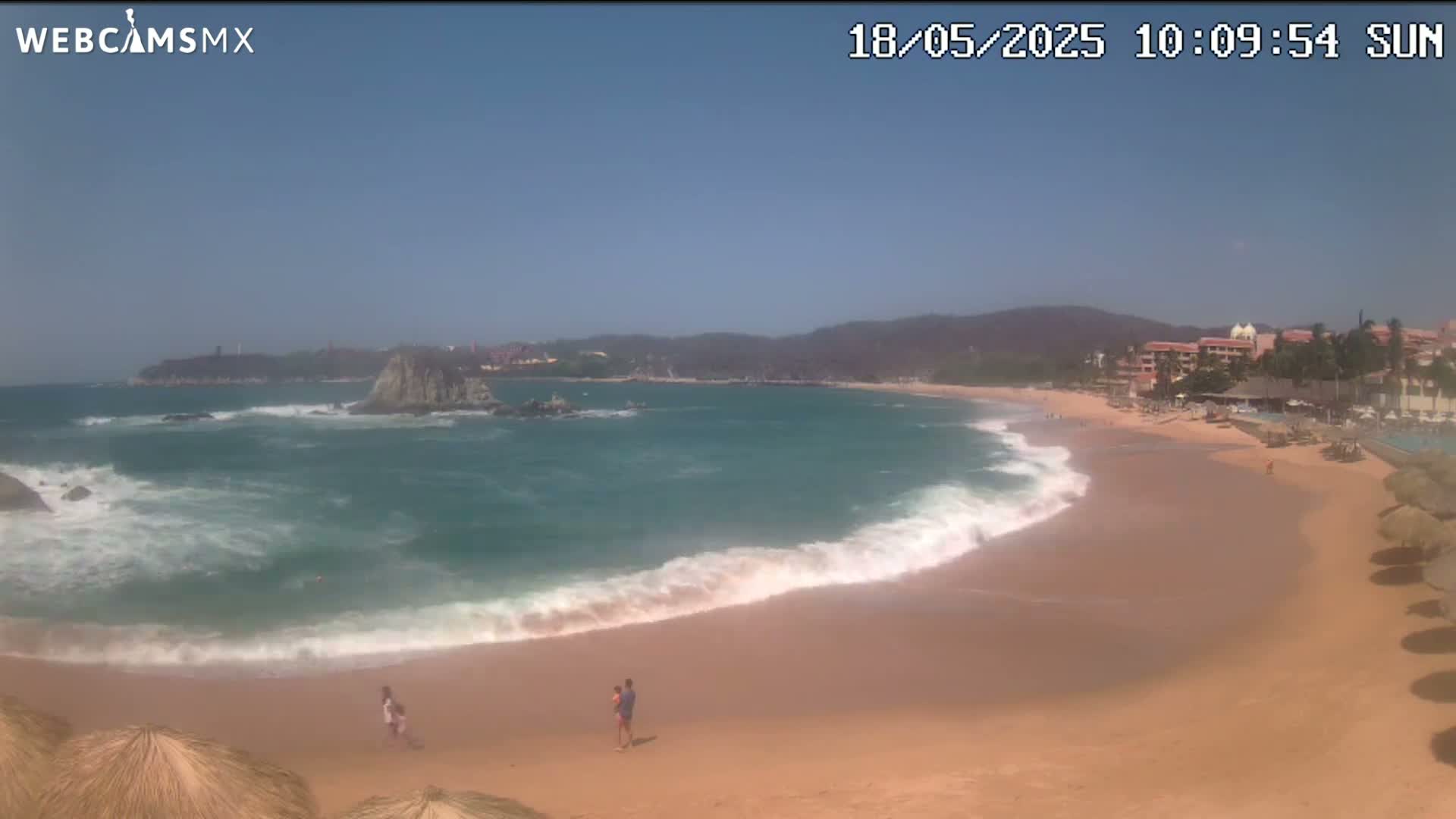 A sandy beach with moderate waves laps the shore under a clear, sunny sky, with a few people visible on the beach and a resort visible in the background.