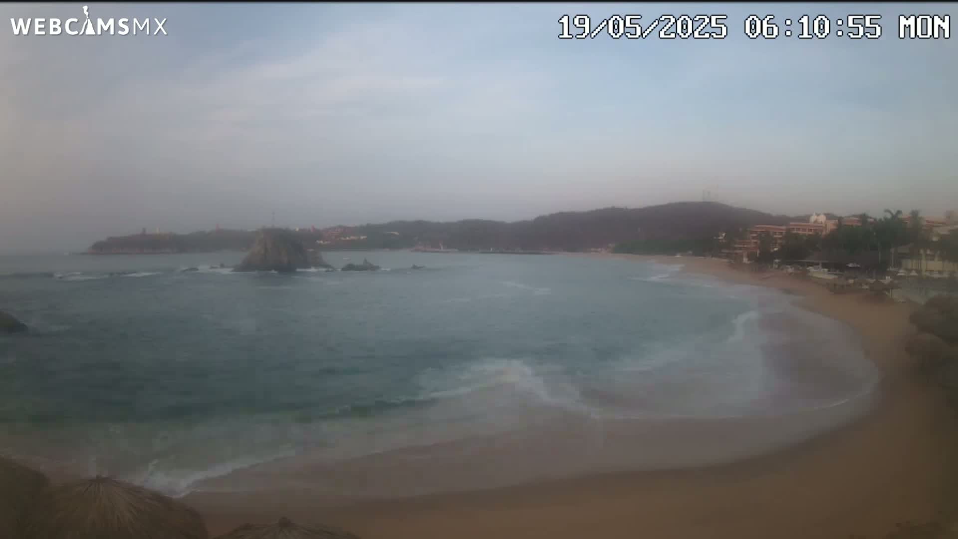 A hazy, overcast morning reveals a sandy beach with gentle waves lapping the shore, backed by a line of buildings and a rocky headland jutting into the ocean.