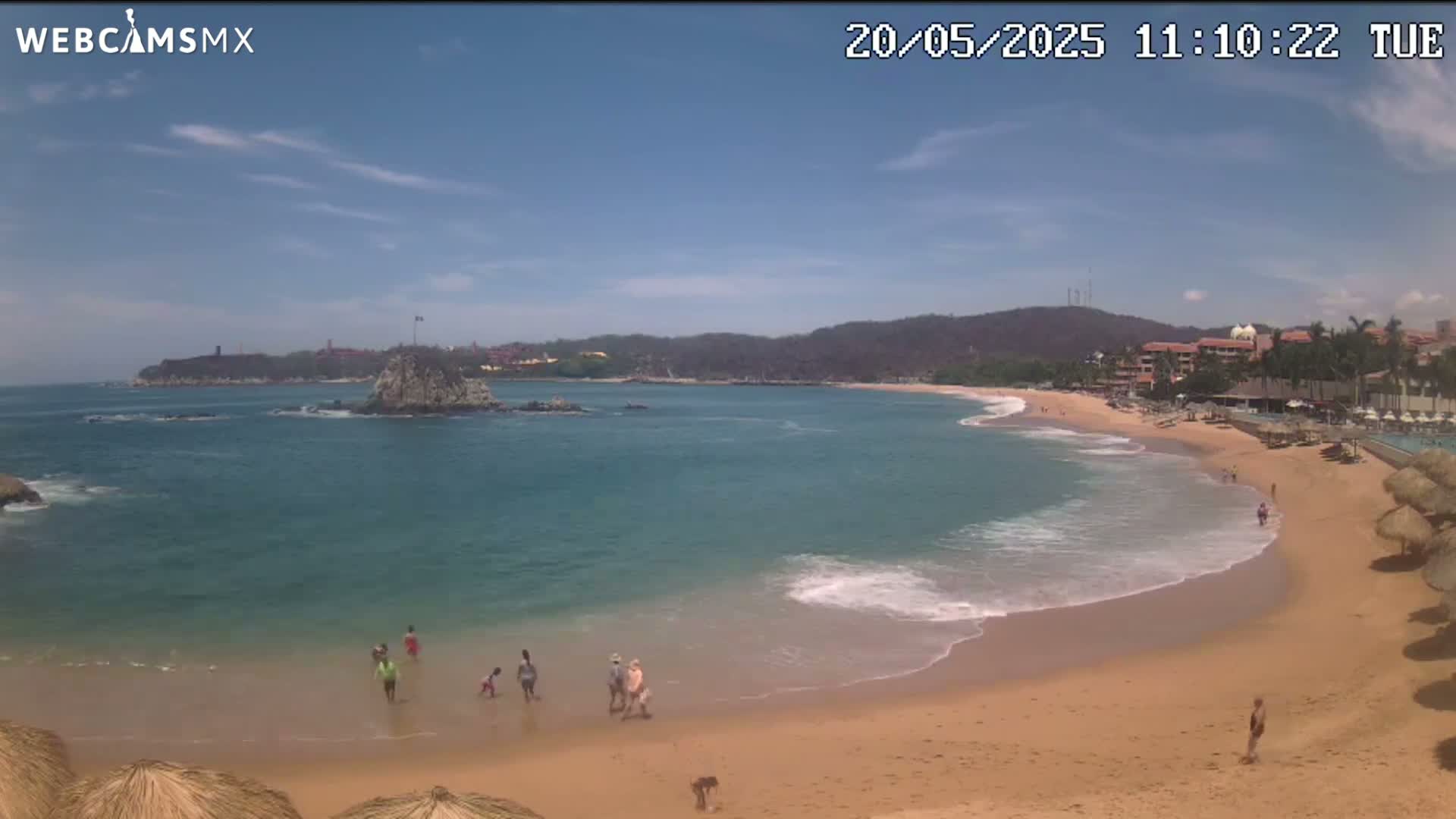 A partly sunny day shows a sandy beach curving around a calm bay with turquoise water, a rocky islet, and distant hills under a mostly clear sky.