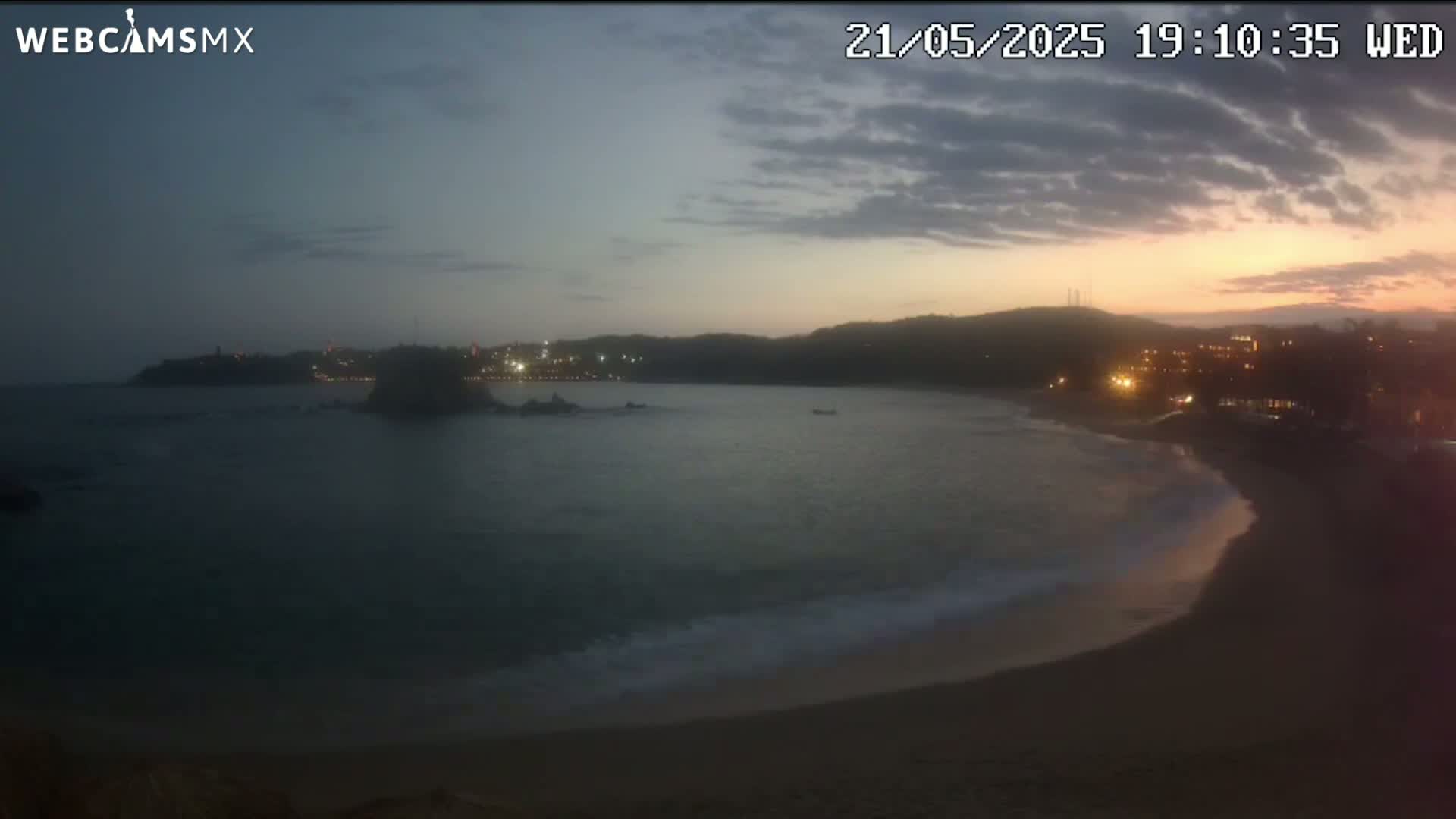 A beach at sunset or twilight shows a calm bay with gently lapping waves, a sandy shore, and distant lights on a hill.