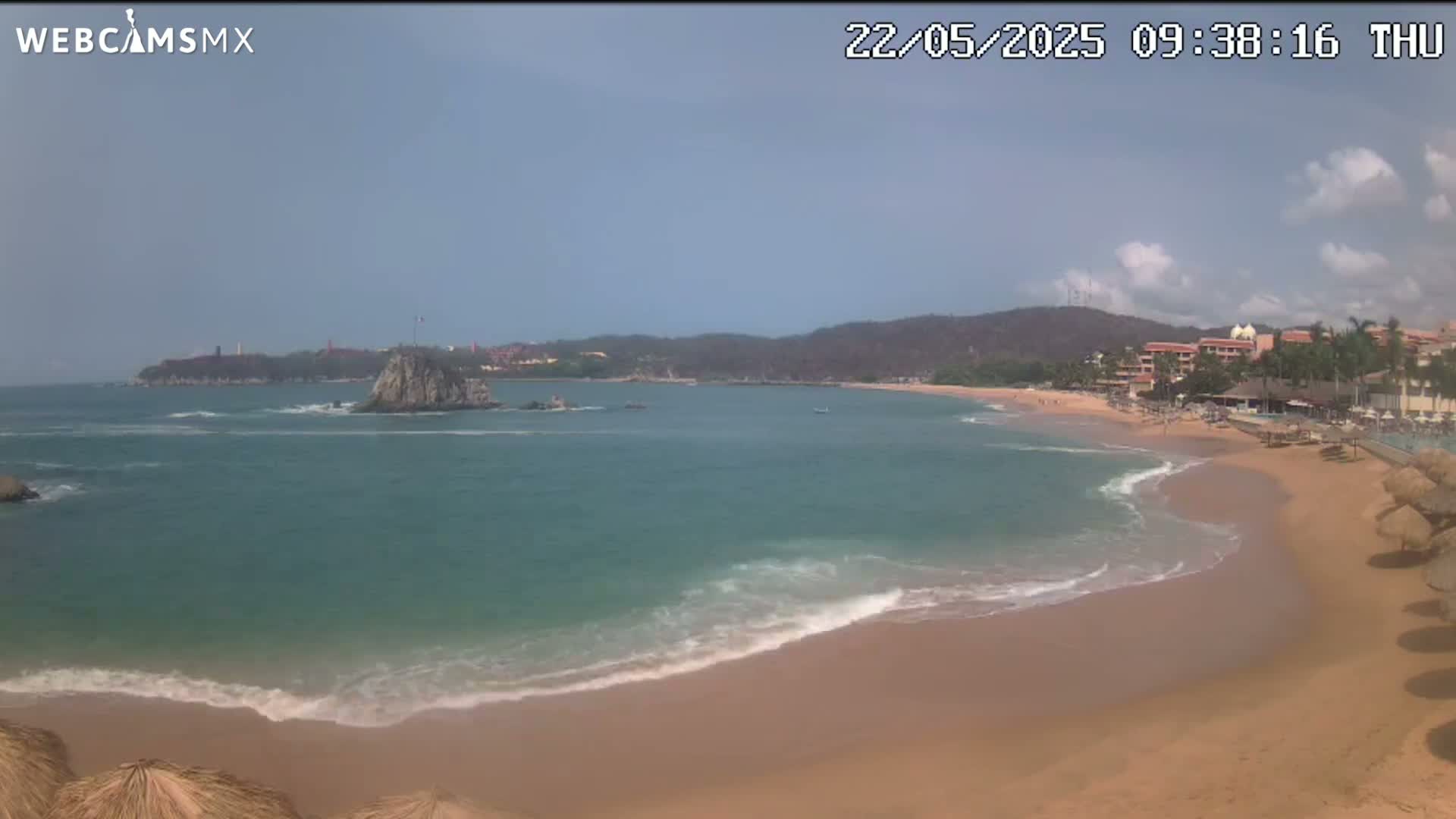 A sandy beach with calm, light-blue water laps against the shore under a partly cloudy sky.