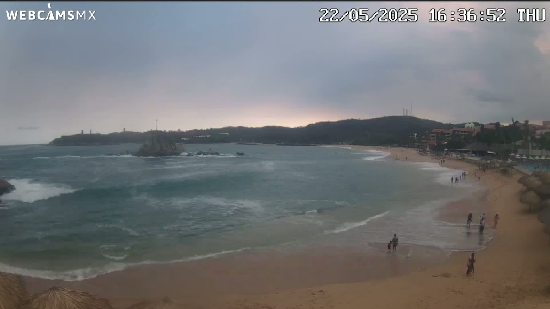 A sandy beach with people walking along the shore and in the ocean, under an overcast sky, with waves lapping the shore and rocky outcrops in the distance.