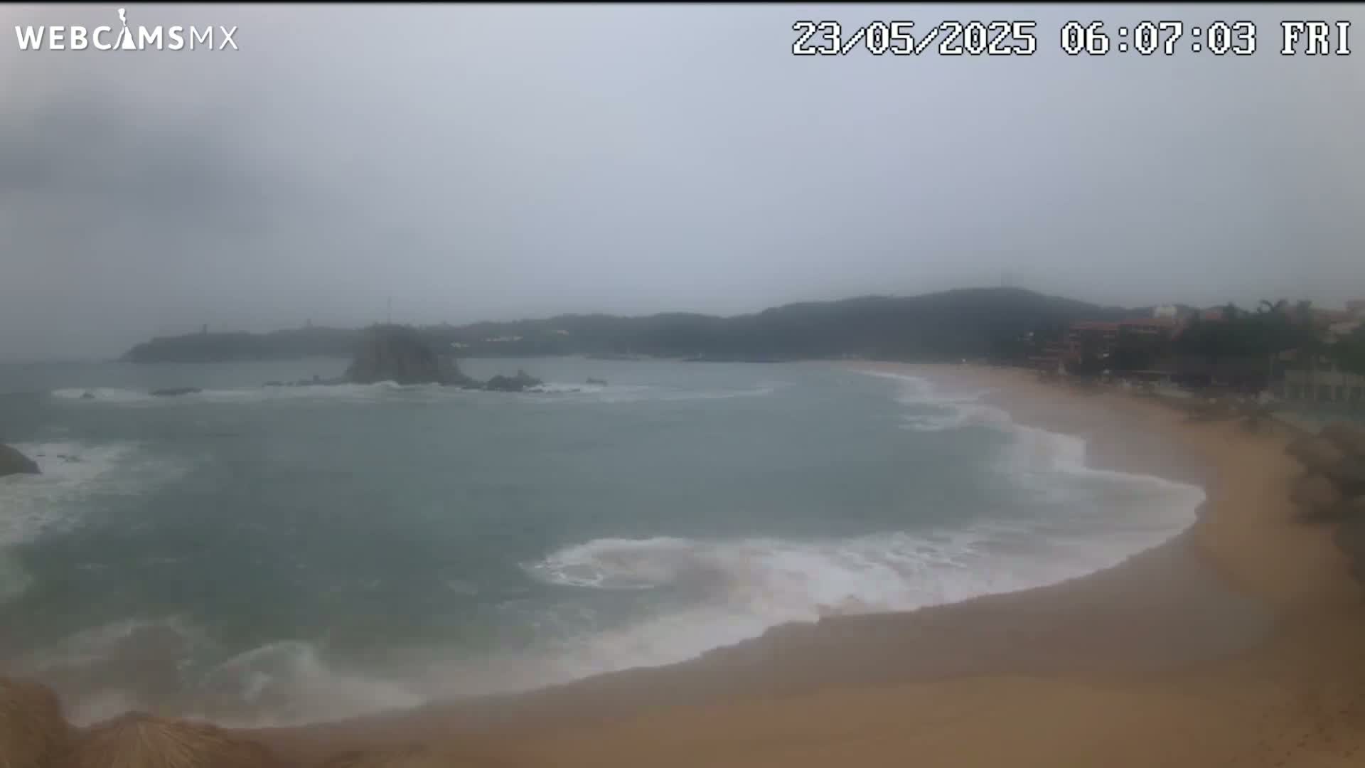A sandy beach with moderate waves laps at the shore under an overcast, hazy sky.