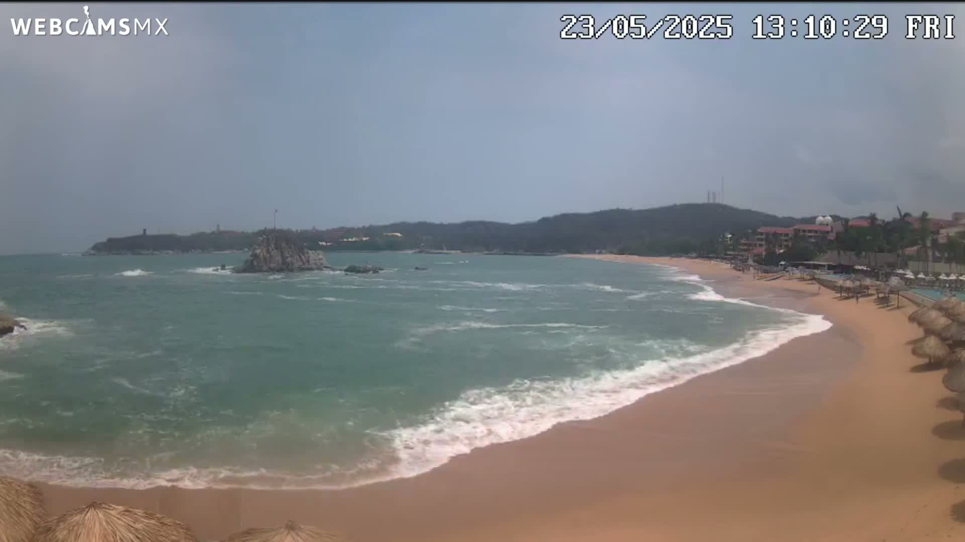 A sandy beach with moderate waves laps the shore under a hazy sky, with a rocky outcropping and distant hills visible across the calm ocean.