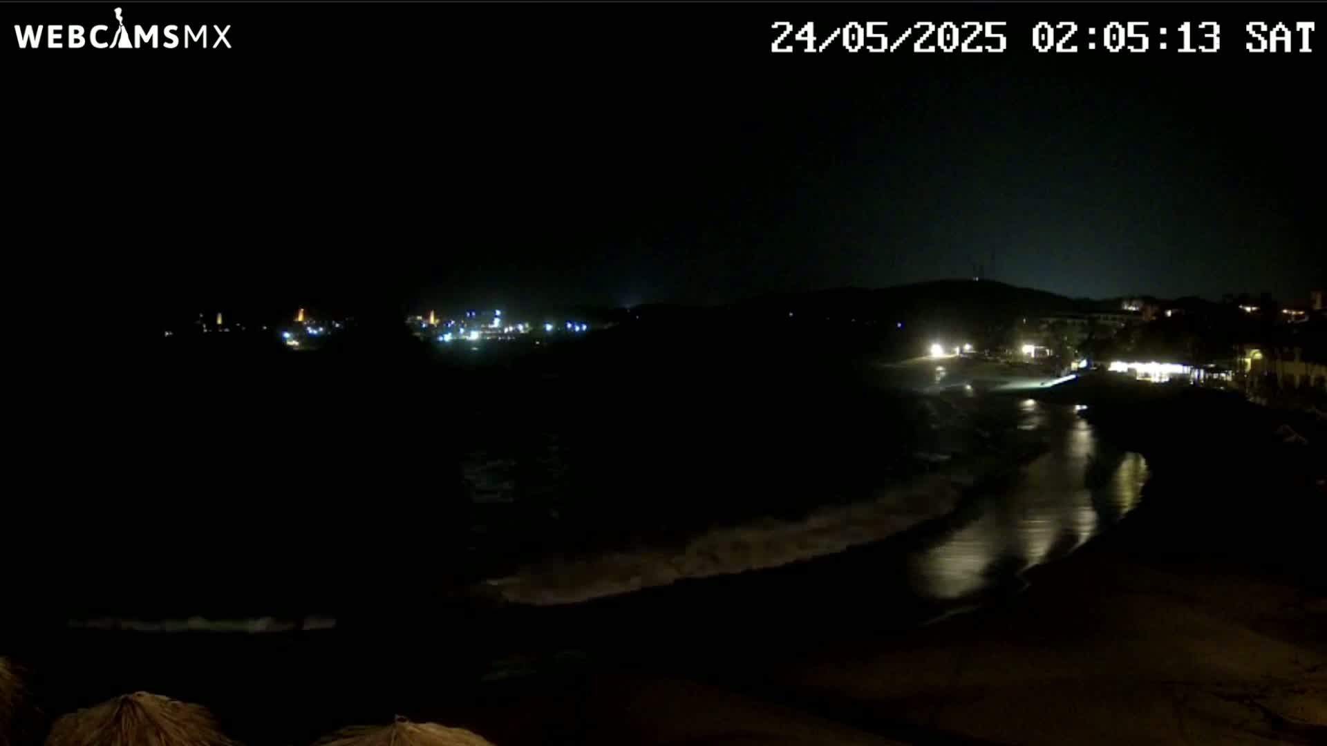 A dark night scene shows a beach with gently lapping waves under a clear sky, and distant lights illuminate a town across the water.