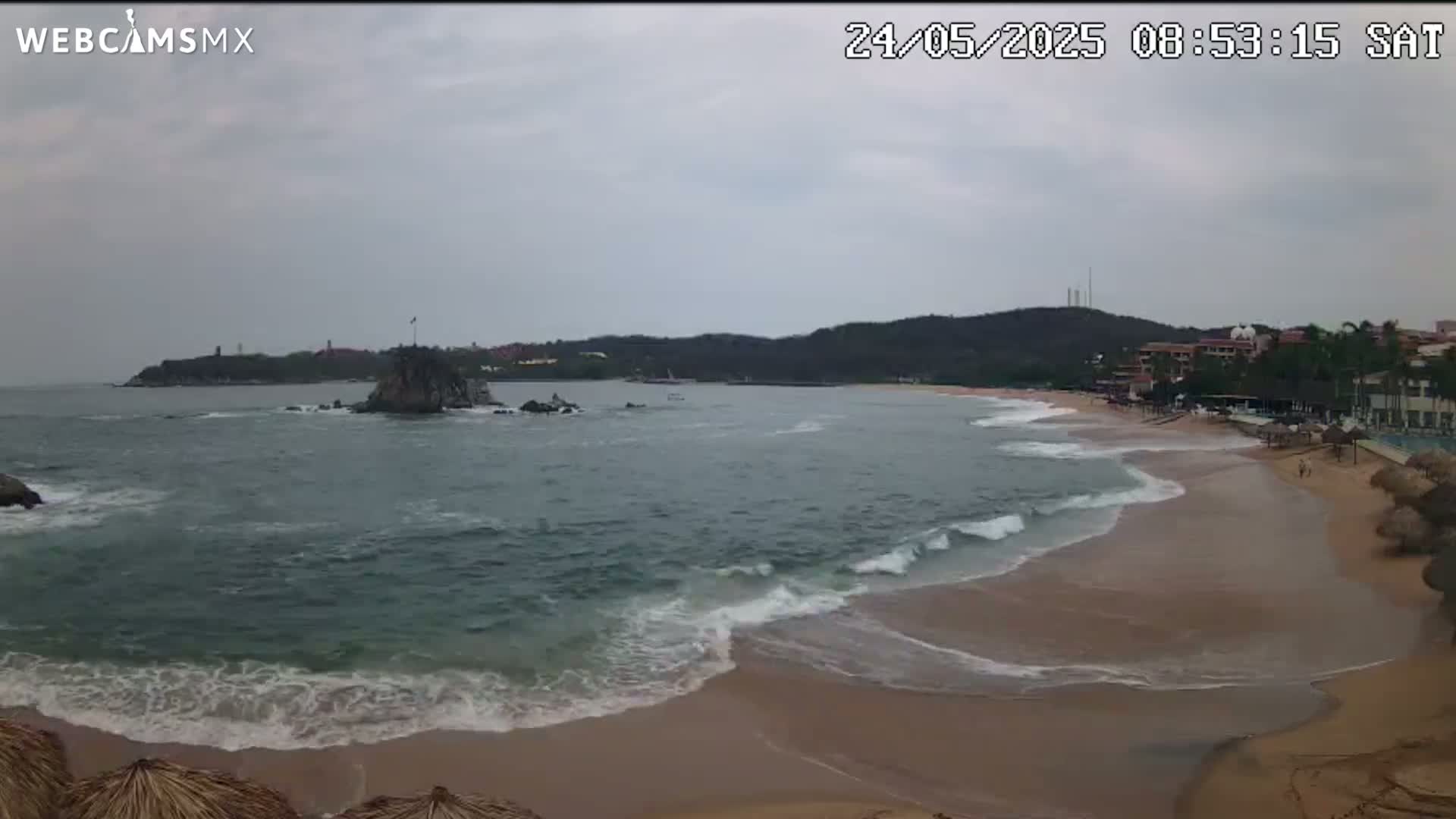 A sandy beach with gentle waves rolls onto the shore under an overcast sky, with a rocky outcrop and distant hills visible across the calm bay.