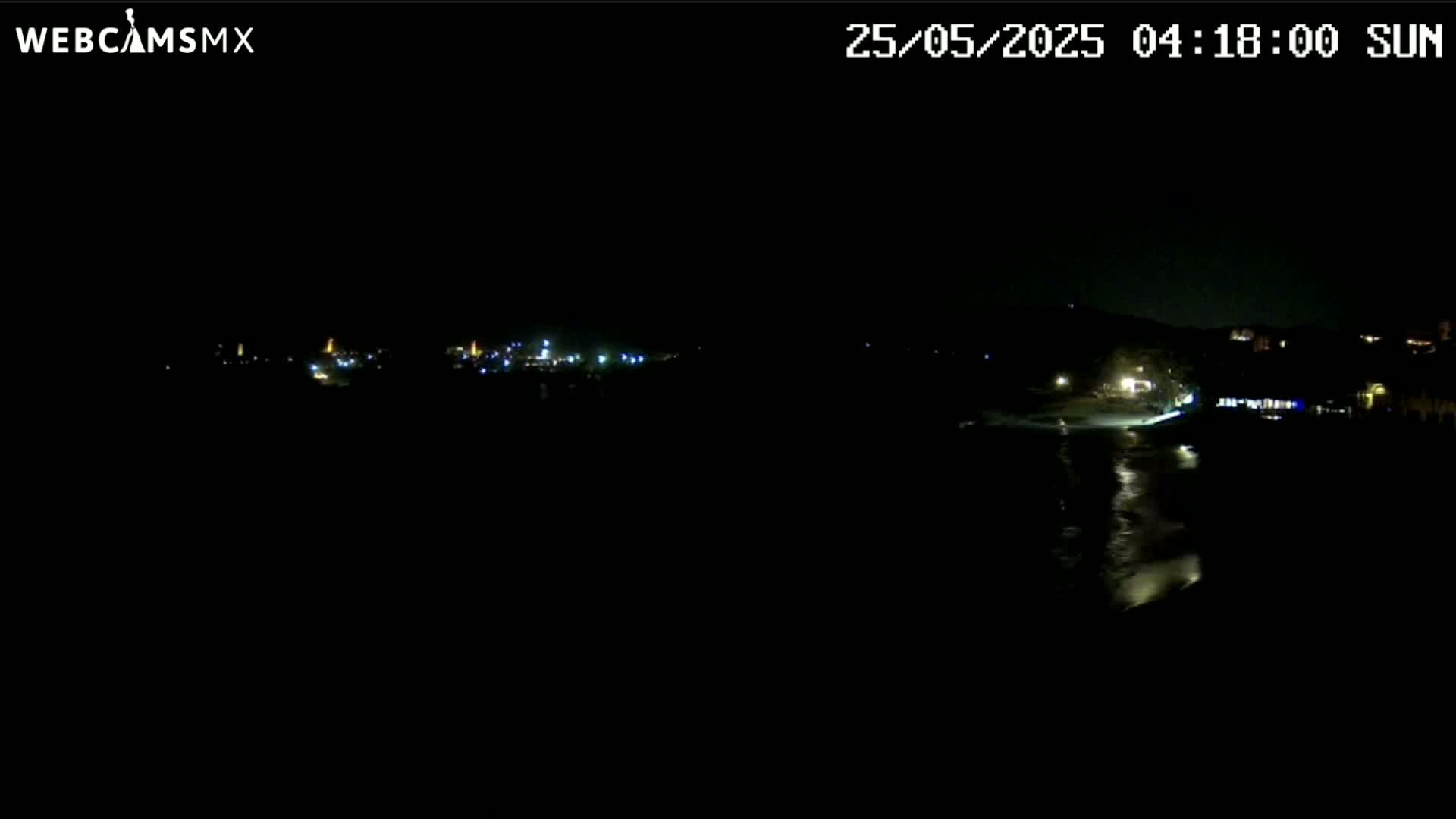 A nighttime long shot shows a dark, mostly obscured shoreline with a few scattered lights from buildings and possibly vehicles.